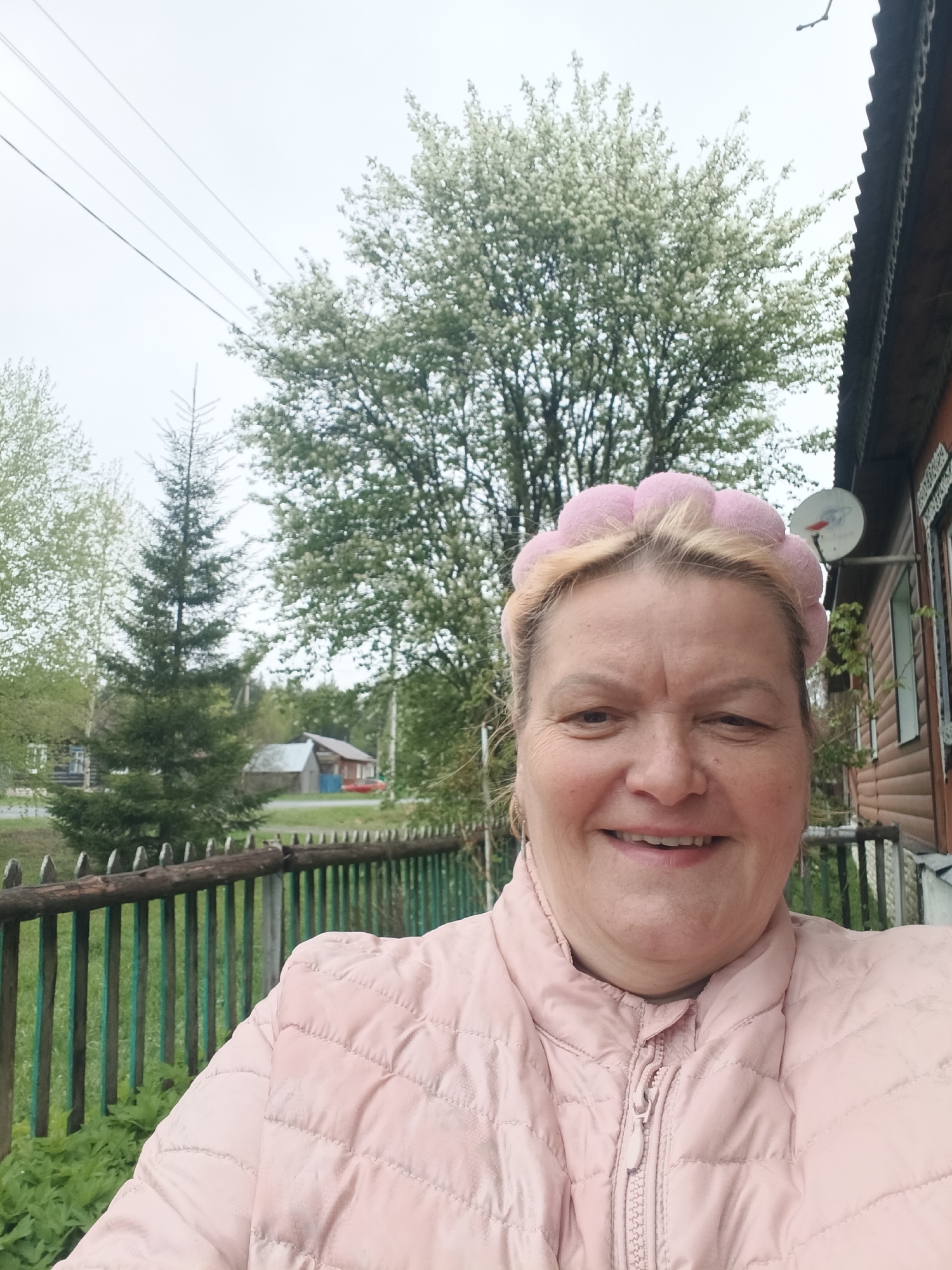 The image shows a middle-aged woman wearing a pink headband and a light pink quilted jacket. She is smiling directly at the camera. Behind her, there is a wooden fence, trees, and a house. A satellite dish is mounted on the side of the house. Power lines are visible in the sky. The location is Красный Маяк, Russia.