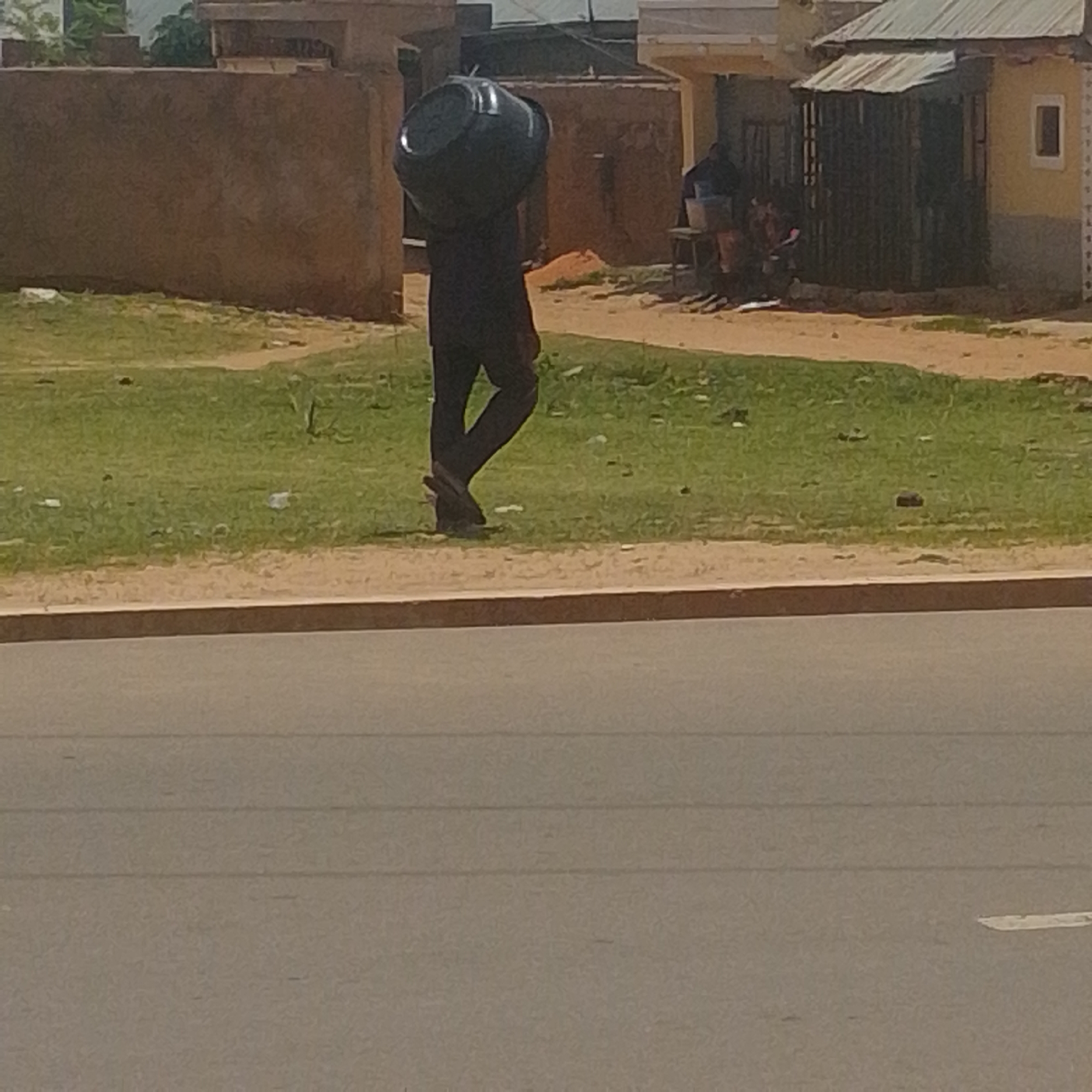 The image depicts a person walking on a dirt path beside a road, carrying a large black plastic bucket on their head. The person is wearing dark clothing. In the background, there are buildings with brown and yellow walls, some with corrugated metal roofs. There is also a grassy area and some trees. The overall setting appears to be a semi-urban or rural environment. The image does not contain any information that allows for the specific location to be identified as Tempure, Nigeria.