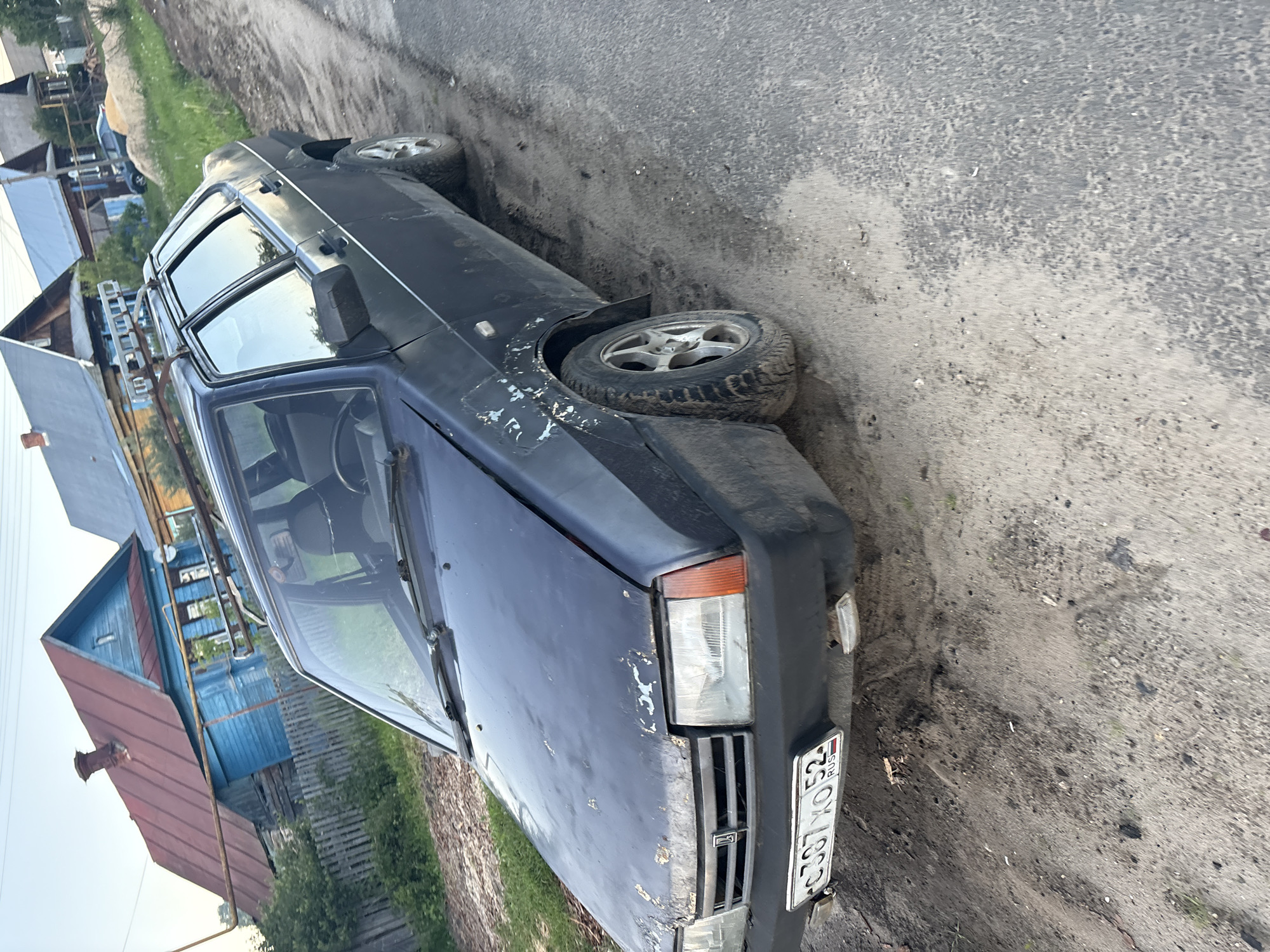 An old, dark-colored car, possibly dark blue or black, is parked sideways on what appears to be a rural or semi-rural road during the daytime. The image is oriented with the car facing towards the top right.
The primary subject is the car, which exhibits significant wear and tear, including peeling paint, scratches, and rust spots, particularly on the front fender and hood. The car features a distinctive front grille with vertical slats and rectangular headlights. It has aftermarket silver alloy wheels, with one visible on the front right and another partially seen on the rear left. The tires appear rugged, with the front right one covered in mud or dirt. A simple, ladder-like roof rack is mounted on the top.
In the background, there are several wooden houses, typical of rural architecture. One house visible to the left has blue walls and a reddish-brown roof. The road surface is a mix of rough asphalt and dirt, with patches of mud and scattered debris. Green grass and some sparse vegetation are visible along the roadside.
No people are present in the image, and there is no discernible activity taking place other than the car being parked. The lighting suggests it is daytime, but the specific time of day is not clearly indicated.
A Russian license plate is clearly visible on the front of the car, displaying the text "C397 XO 52 RUS." The "52 RUS" code indicates the Nizhny Novgorod Oblast region of Russia, providing a strong geographical location cue. The overall scene suggests an older, utilitarian vehicle in a modest, perhaps weathered, local setting.