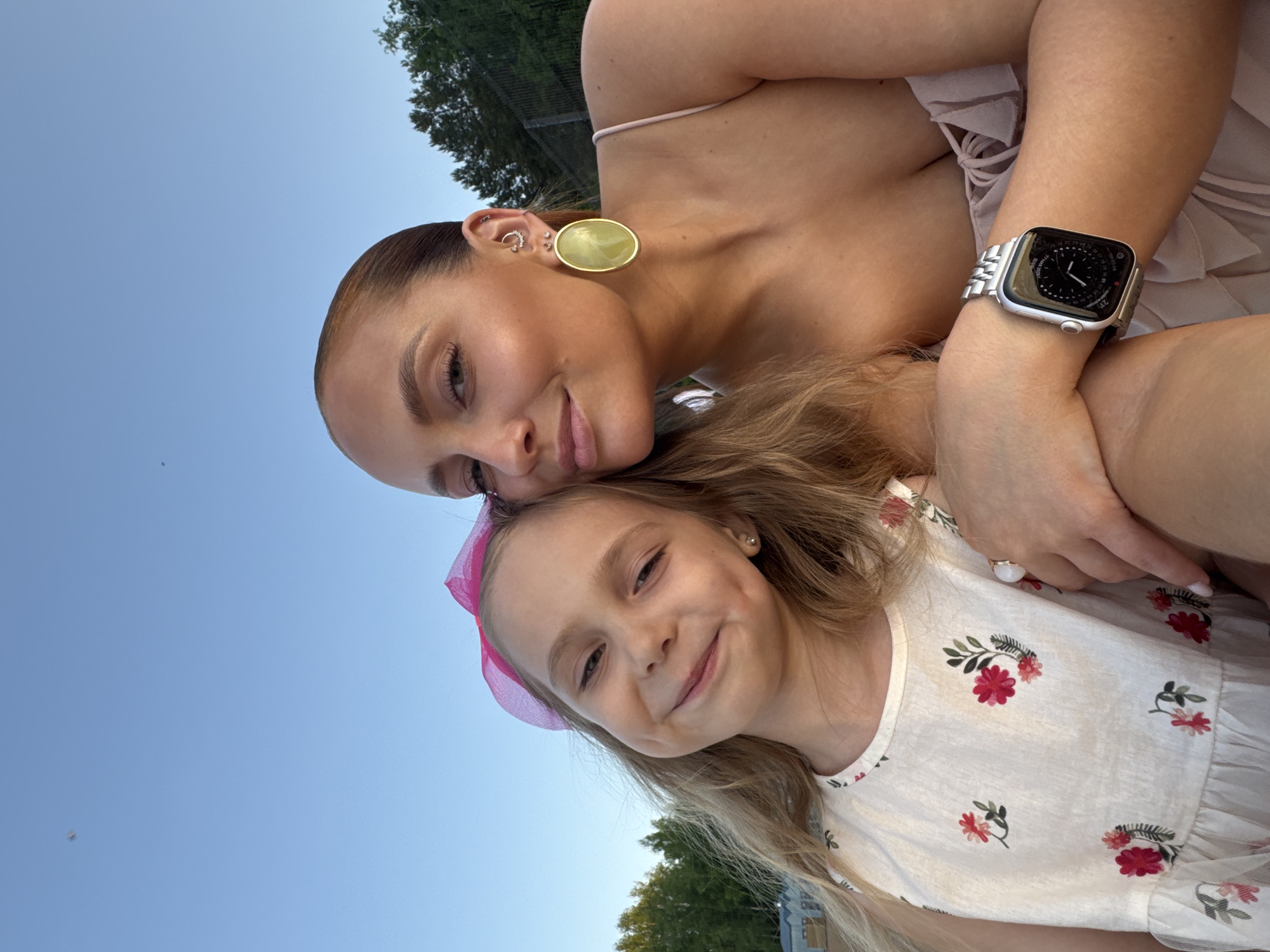 A woman and a child pose together in an outdoor setting. The woman has fair skin, brown hair pulled back, wearing large gold earrings and a silver smartwatch. The child, presumably a daughter, has blonde hair, a pink bow, and is wearing a white dress with red floral patterns. The background suggests a park or garden, with trees and a clear sky. The photograph was taken in the city of Chaikovsky, Russia.