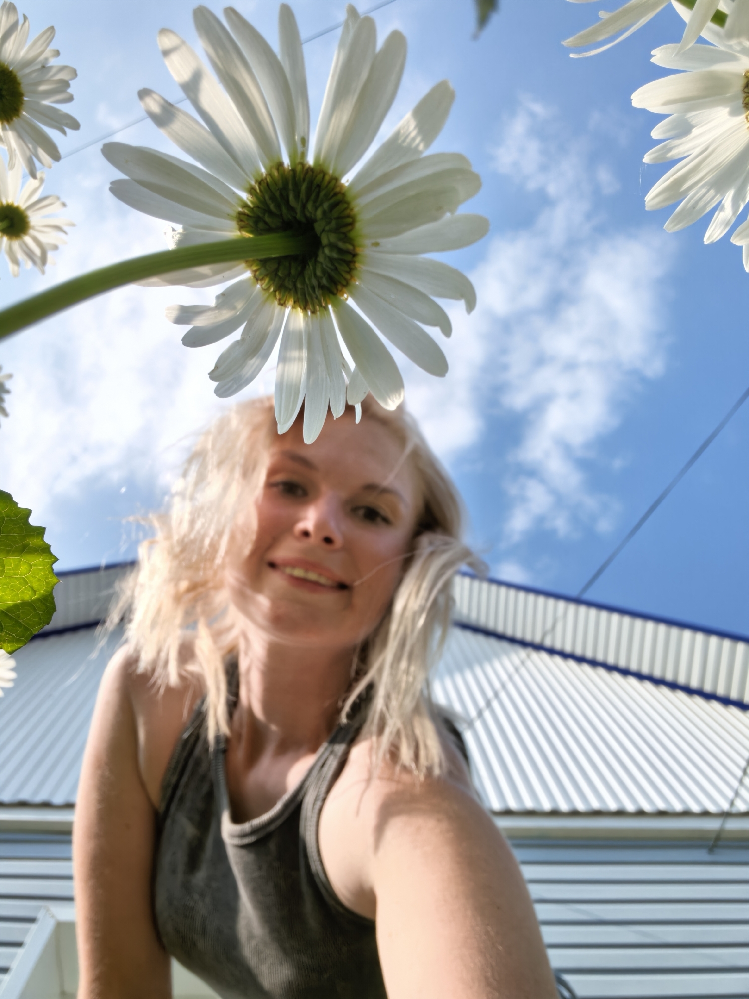 The image, taken from a low-angle perspective, features a woman with blonde hair smiling and looking towards the camera. She is wearing a dark tank top. Daisies, viewed from underneath, dominate the foreground with their white petals and green centers, set against a bright blue sky with scattered clouds. In the background, a white corrugated roof of a building is visible. The scene appears to be outdoors, with a wire running across the upper right portion of the frame. The location is identified as Sterlitamak, Russia.