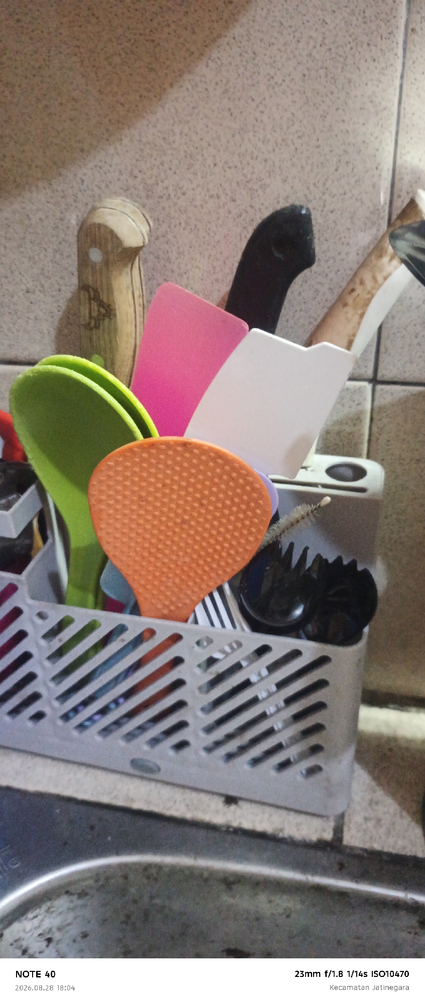 The image shows a close-up, eye-level, indoor shot of kitchen utensils in a grey plastic holder on a tiled backsplash. A wooden-handled knife with a black blade is visible, along with a pink spatula, a white spatula, and a bright green spoon. An orange, heart-shaped strainer with raised dots is also prominent, as are a few black utensils with serrated edges. The background is a speckled, light brown tiled wall. The bottom of the image shows a stainless steel sink with some debris and the text "NOTE 40 2026.08.28 18:04 23mm f/1.8 1/14s ISO10470 Kecamatan Jatinegara". The location is identified as Kecamatan Jatinegara, East Jakarta, Indonesia.