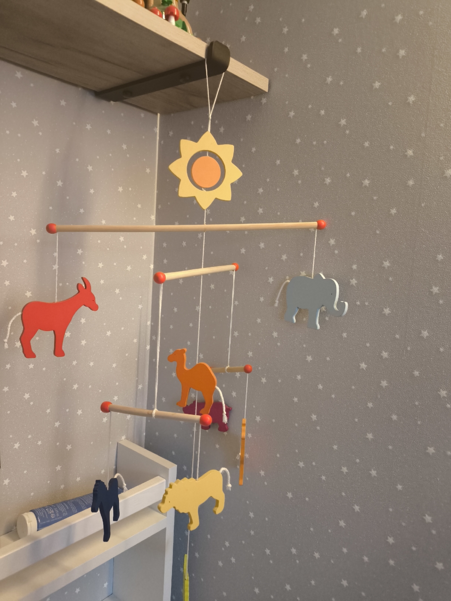 The image depicts a baby mobile hanging in a room with star-patterned wallpaper. The mobile features a yellow sun at the top, followed by horizontal wooden bars with red ends from which various wooden animal shapes are suspended: a red donkey, a gray elephant, an orange camel, a blue bird or bat-like figure, and a yellow lion. The mobile is attached to a shelf above. A tube is visible on the shelf below. There is no evidence to support that this is in the city of Moscow, Russia.