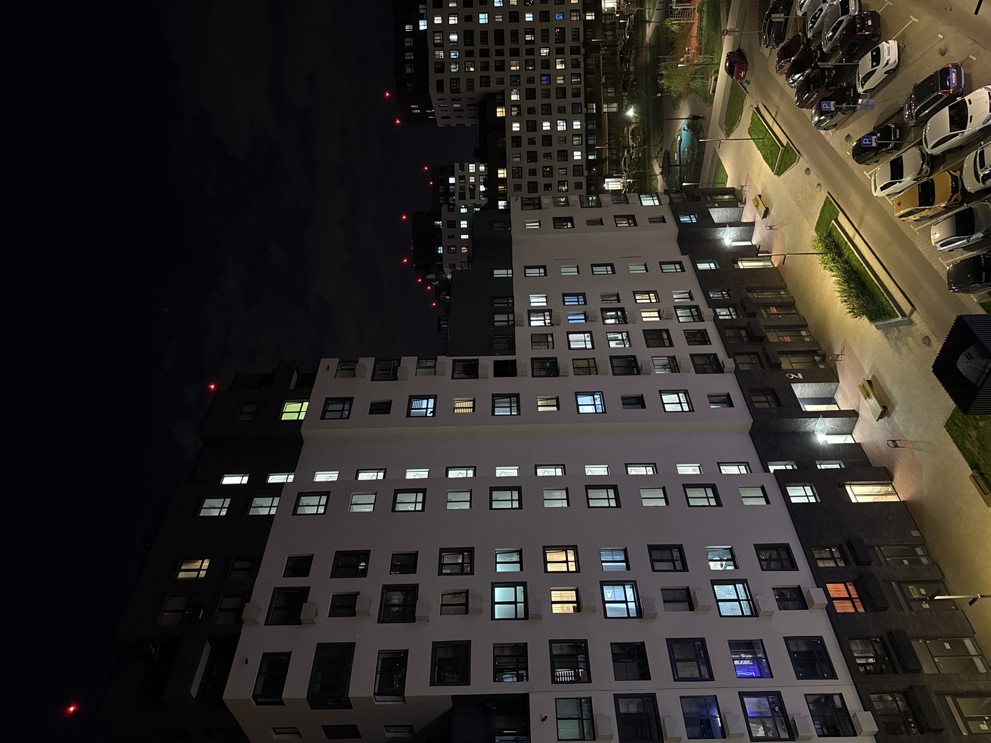 This is a nighttime, eye-level shot of residential buildings and a parking lot in Troitsk, Russia. The buildings are tall, modern structures with numerous windows, some of which are illuminated. One building has a stark contrast between a white façade and a dark grey one. A parking lot is seen beside one of the buildings, filled with various cars. The sky is dark and the overall atmosphere is urban.