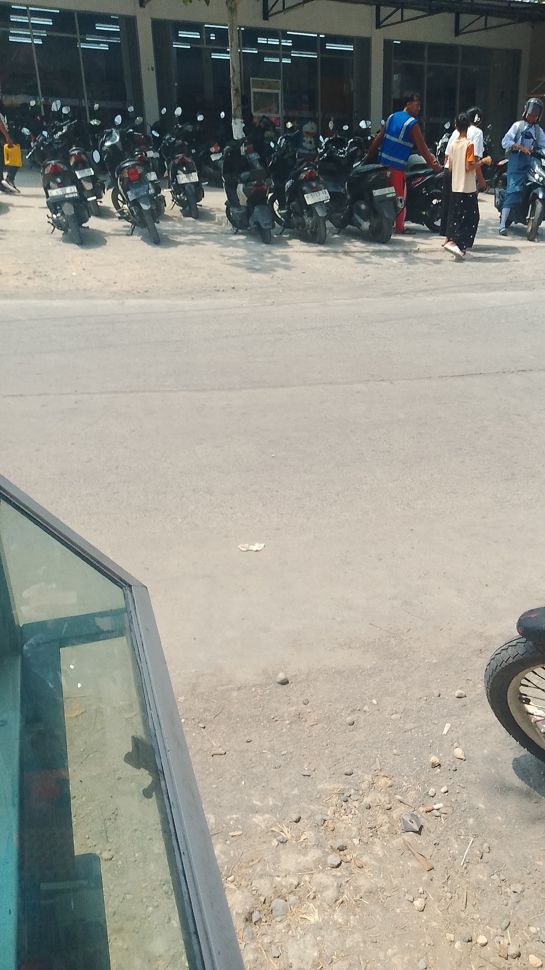 In Karanglangu, Indonesia, a row of parked motorcycles is visible on a dusty, unpaved surface adjacent to a building with large glass windows. Several individuals are present. One person, wearing a blue vest and red pants, appears to be interacting with a motorcycle. Another individual, dressed in a light-colored shirt and dark patterned skirt, walks away from the motorcycles. A third person, in a light blue uniform and helmet, sits on a motorcycle. The ground is littered with small rocks and debris. In the foreground, a portion of a glass structure with a dark frame is visible.