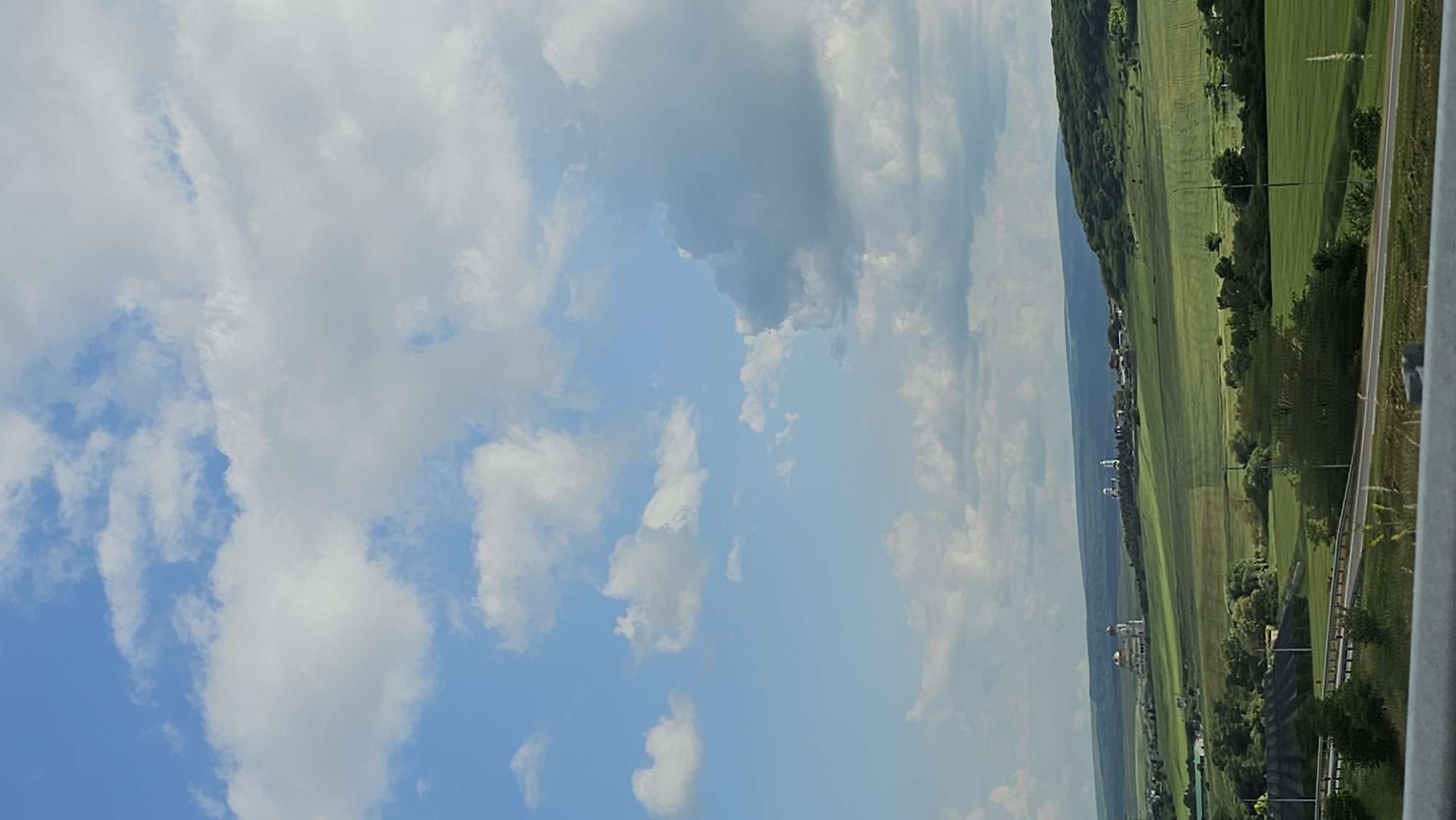 This media file is a vertical photograph depicting a landscape scene under a partially cloudy sky. The primary subjects are the sky, a green, rolling landscape with a road, and some distant buildings.
The scene appears to be a rural or semi-rural area with vast fields and rolling hills, characteristic of a countryside setting. A road runs along the right side of the image, separating the viewer from the expansive green fields. The fields themselves are neatly divided, suggesting agricultural activity. In the distance, several buildings are visible, including structures that appear to be churches or other significant architectural elements, some with golden domes. The overall setting suggests a serene and picturesque view, possibly taken from a highway or an elevated vantage point.
The sky is a prominent feature, with a mix of bright blue patches and large, fluffy white clouds, indicative of fair weather with some cloud cover. The presence of these clouds suggests it might be daytime, likely midday or afternoon, given the brightness of the scene. There is no discernible activity or event taking place; it is a still landscape shot.
Notable details include the distinct green hues of the fields, the texture of the clouds, and the architectural style of the distant buildings. The road has white lane markings. The overall mood is peaceful and tranquil, offering a glimpse of rural beauty. There is no visible text in the image. The location context provided indicates Yessentuki, Russia, which aligns with the visual elements of churches and the type of landscape.