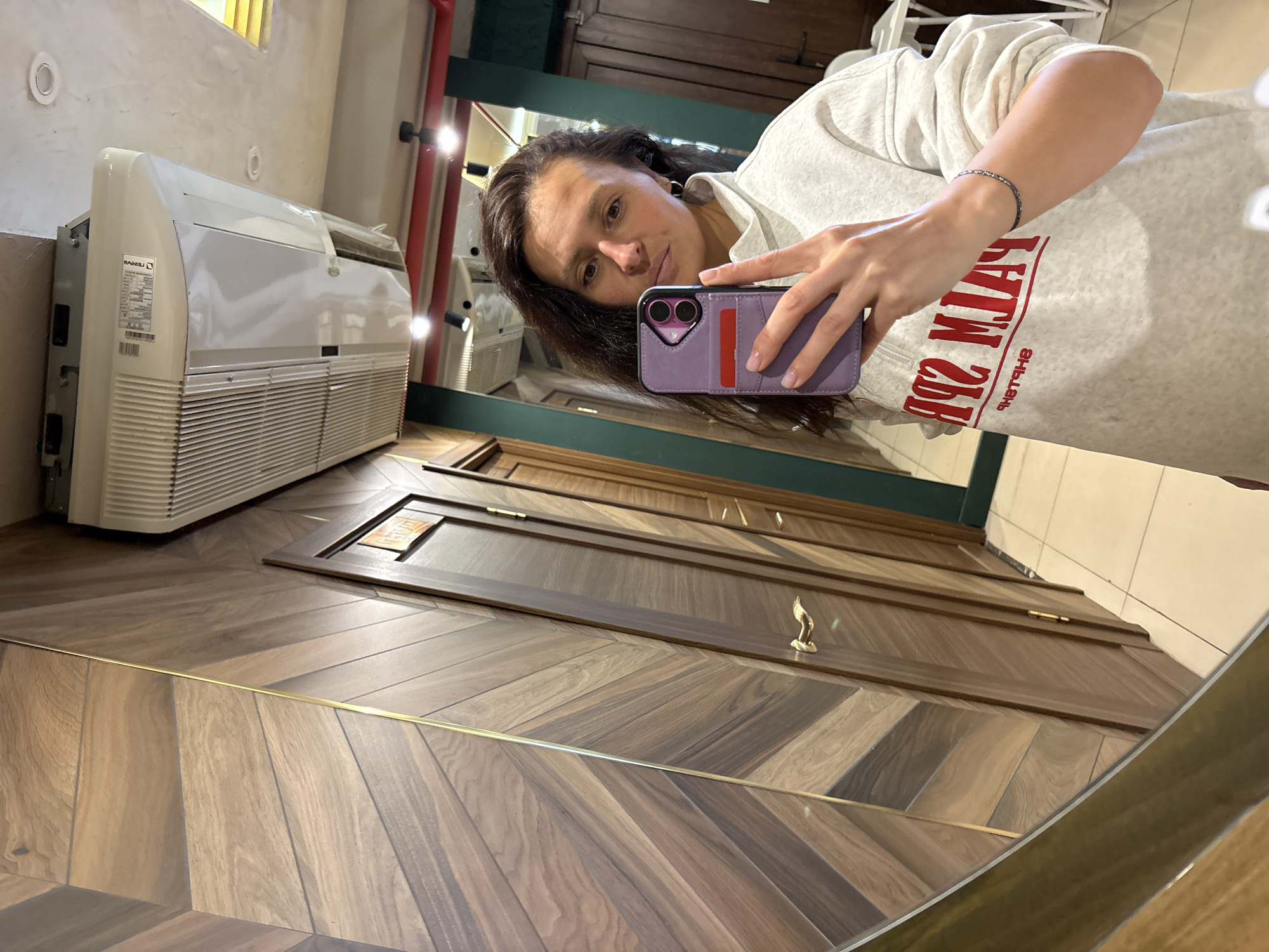 The image depicts a woman taking a selfie in a mirror. The mirror reflects a room with a herringbone wood floor and a white air conditioning unit mounted on the wall. The woman is holding a purple smartphone with a wallet case. The reflection includes the air conditioning unit, wood flooring, a dark green door, and a red column. There is insufficient information to determine the location within Neftekamsk, Russia.
