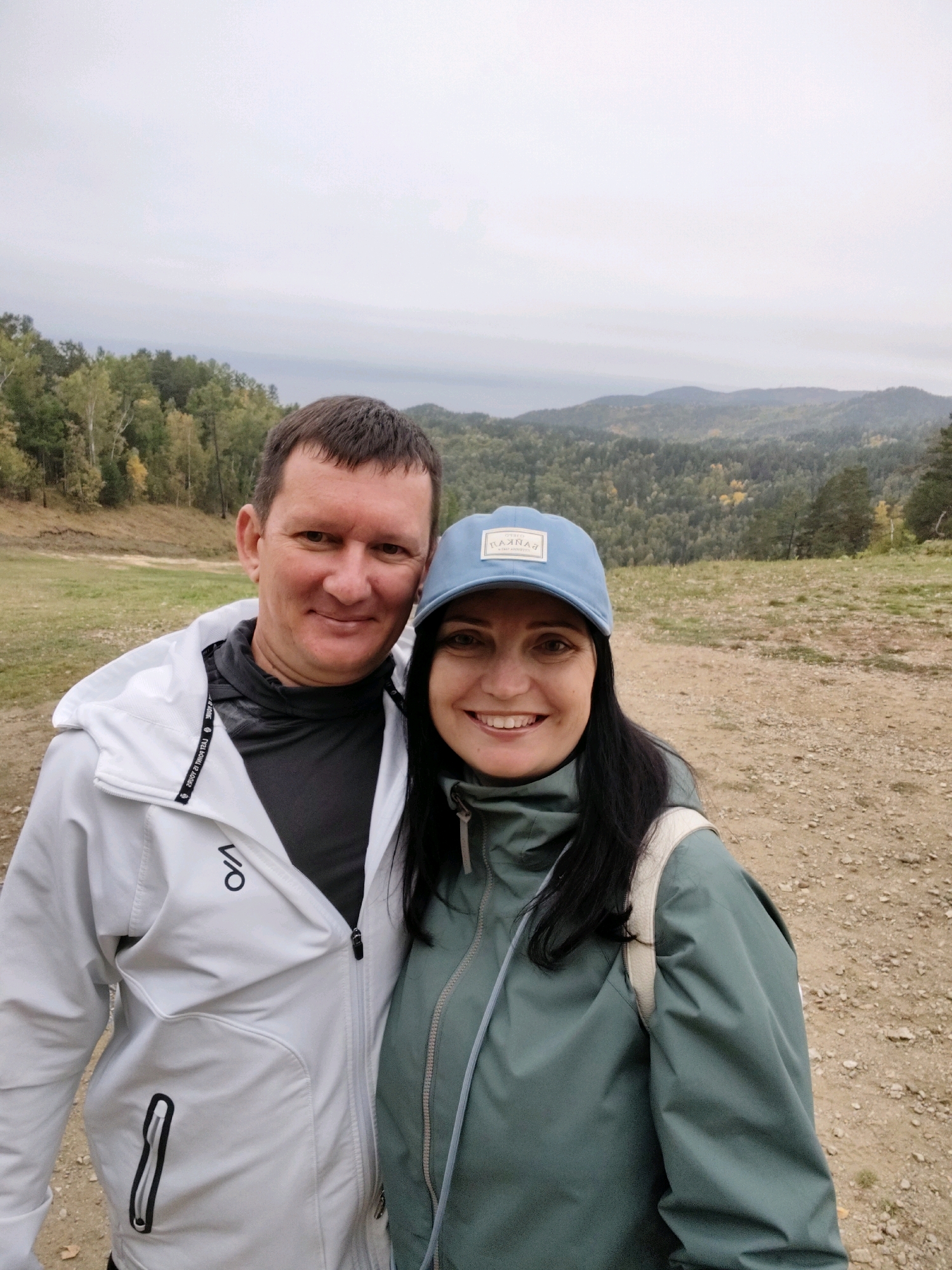 A man and a woman are posing for a selfie in an outdoor environment. The man is on the left, wearing a white zip-up jacket over a dark gray hooded shirt. He has short brown hair and is smiling. The woman is on the right, wearing a green hooded jacket and a light blue baseball cap. She has dark hair and is also smiling. They are standing close together, with the man's arm around the woman's shoulders. Behind them, there is a landscape of rolling hills covered in trees. The sky is overcast. The ground in the foreground is a mixture of dirt and small stones. The location is identified as Nikola, Russia.
