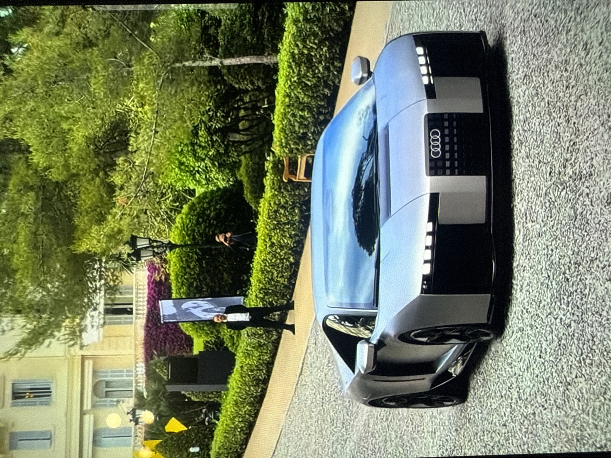 This image depicts an outdoor automotive display, specifically featuring a silver Audi concept car, set against a backdrop of a grand building and manicured gardens. The scene is located in the city of Varna, Bulgaria.
In the foreground, a sleek, low-profile, silver-grey Audi concept car is parked on a light-colored, pebbled surface, angled towards the viewer's right. The car features a prominent Audi four-ring logo on a dark, patterned front grille, flanked by linear LED light elements on the bumper and above. Its windshield reflects the sky and surrounding greenery.
In the mid-ground, on a paved path lined by green hedges, a man in a dark suit with a white shirt stands facing the camera, hands behind his back. To his right, partially obscured, another person in dark attire holds a camera, actively capturing the scene.
The background is dominated by a large, ornate, light-colored building with multiple windows, some fitted with open and closed shutters, and balconies adorned with balustrades. Surrounding the path and building are extensive, well-maintained gardens featuring neatly trimmed green hedges, a variety of green trees and bushes, and a section of purple flowering plants. Event elements are visible, including yellow triangular flags, dark banners (one with a white graphic), black speaker units, and two multi-lantern black lampposts. A wooden bench is situated behind a hedge section on the right. The scene is illuminated by natural daylight.