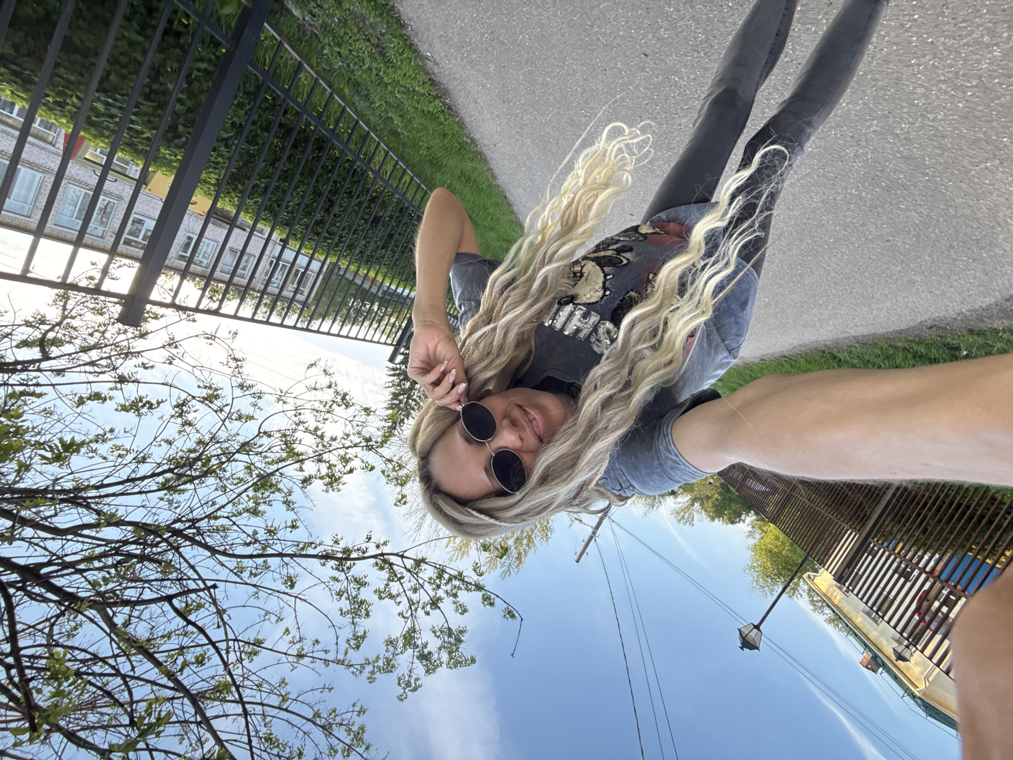 A woman stands on a paved path, likely in the city of Rusanovo, Russia, indicated by the presence of buildings typical of the region. She is wearing sunglasses, a t-shirt, and jeans. A metal fence separates the path from a grassy area. Trees and a clear blue sky are visible in the background. The image appears to be a selfie, taken at an unusual angle.