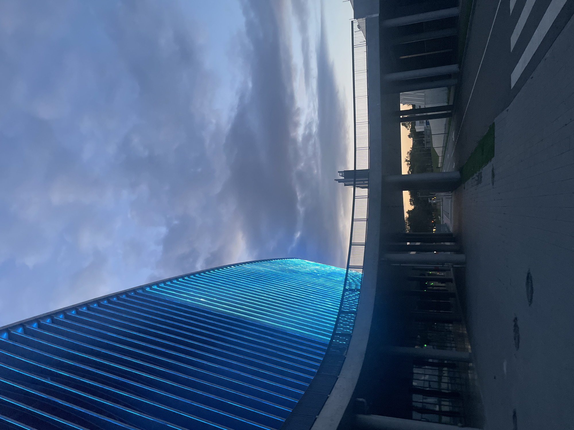 The image depicts the exterior of a modern building with a curved facade illuminated by bright blue lights. The building has a sleek, ribbed design. In the foreground, there is a paved walkway with visible manhole covers. A pedestrian bridge with a metal railing extends across the scene, supported by concrete pillars. Below the bridge, a road is visible with a crosswalk. The sky is cloudy and appears to be twilight, with soft, muted colors. The overall environment suggests an urban setting, likely a public space or entertainment venue, given the architectural style and lighting. There are no people visible in the image.