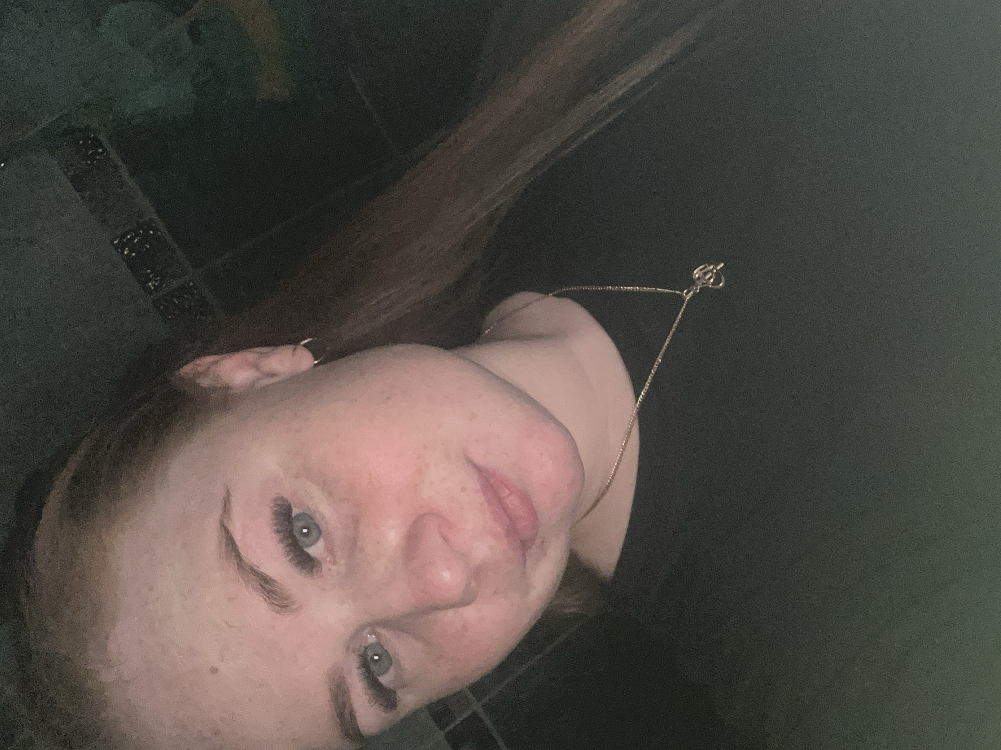 A close-up, dimly lit selfie of a woman with light skin and light brown hair. She has visible freckles, light blue eyes, and extended eyelashes. She is wearing a gold necklace. The background consists of dark, mottled surfaces, possibly a wall or other interior element. No landmarks or features present in the photograph can confirm this was taken in Chaikovsky, Russia.