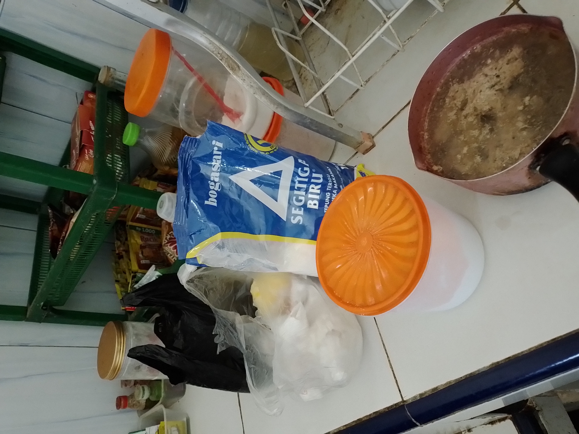 The image shows a kitchen counter with various food items and cooking utensils. In the foreground, a pot filled with a liquid substance, possibly soup or broth, is seen on the right. To the left of the pot, there is a white container with an orange lid, containing a white powdery substance, likely flour. Behind this, a large blue bag labeled "bogassari SEGITIGA BIRU TEPUNG TERIGU SERBAGUNA" (all-purpose wheat flour) is prominent. Next to the flour bag, a clear plastic bag contains white fluffy material, possibly cotton balls or a food ingredient. A black plastic bag is also visible, partially obscuring the other items.
In the background, a green shelf unit holds several packaged food items and bottles. Among them are clear plastic bottles with green and orange caps, containing liquids. There's also a clear jar with a gold lid. The overall environment appears to be a domestic kitchen setting. The presence of "bogassari" brand flour suggests the location is likely Indonesia. Given that Ngancar is a district in Kediri Regency, East Java, Indonesia, it is plausible this kitchen is located there.