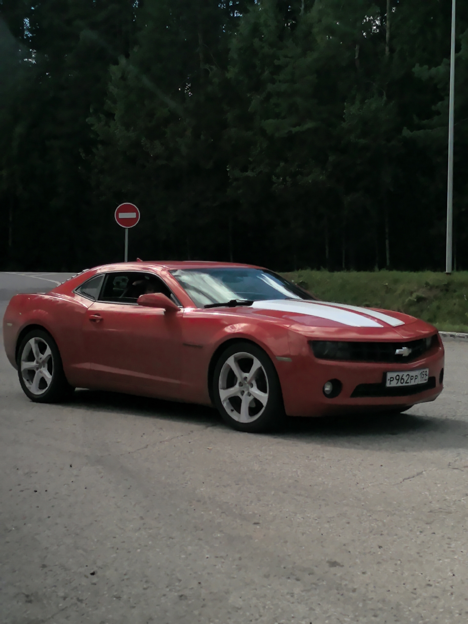 A red Chevrolet Camaro with white racing stripes on the hood is parked on a paved road. The car is viewed from the side at an angle, showing its front and passenger side. A license plate with the characters "P962PP 159" is visible on the front bumper. In the background, a dense forest of evergreen trees forms a dark backdrop. A red and white circular traffic sign with a horizontal white bar indicates a "no entry" restriction. The sky is not clearly visible, suggesting it might be late evening or overcast. The overall scene suggests a car parked on the side of a road, possibly a highway or a scenic route, adjacent to a forested area. The specific location in the city of Vary, Russia, cannot be determined from the visual information alone.