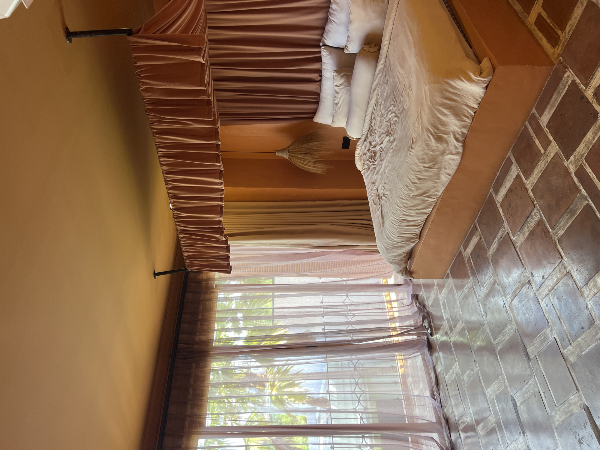 The image depicts a bedroom in North Kuta, Indonesia, featuring a large bed with light-colored bedding and several pillows. The room has an orange-toned ceiling and walls. Drapes in a coordinating orange hue hang from the ceiling above the bed, creating a canopy effect. A woven pendant light hangs near the head of the bed. Large windows covered by sheer, pinkish curtains offer a view of palm trees and a balcony railing. The floor is covered with terracotta-colored tiles laid in a brick pattern. The overall ambiance suggests a tropical, resort-style setting.