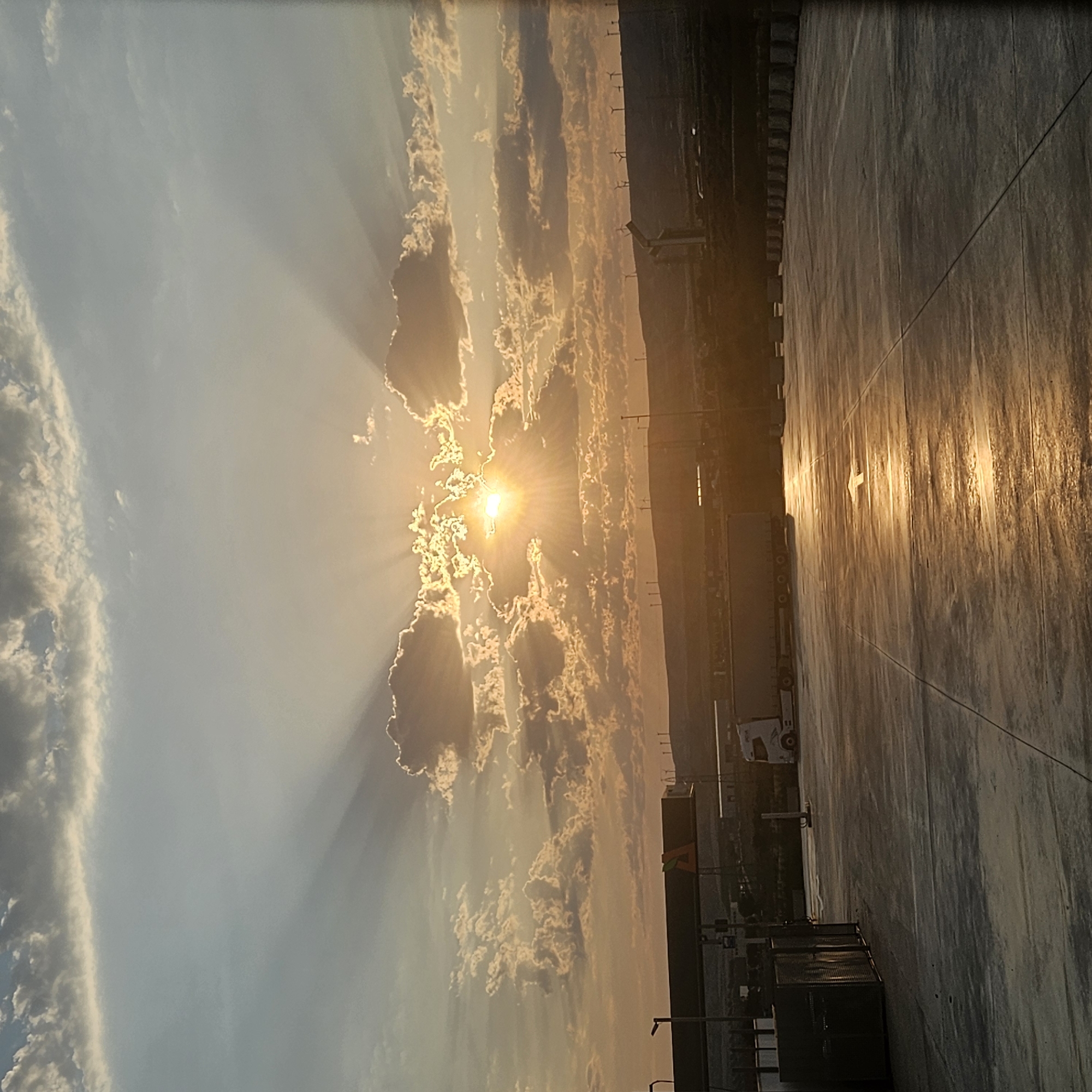 The image depicts a sunset over a commercial or industrial area, possibly a truck stop or rest area, in Épila, Spain. The sun is low on the horizon, peeking through dramatic, illuminated clouds, casting long rays of light across the sky. The foreground is a paved surface, likely concrete, reflecting the warm hues of the sunset and showing a painted white arrow indicating direction. To the left, a structure resembling a bus shelter or covered waiting area is visible, along with some large, dark rectangular objects. Further into the midground, a white semi-trailer truck is parked. In the background, a low, rolling hill is lined with wind turbines, characteristic of the region's renewable energy infrastructure. The overall atmosphere is one of a quiet end to the day, with the natural beauty of the sunset contrasting with the man-made structures.
