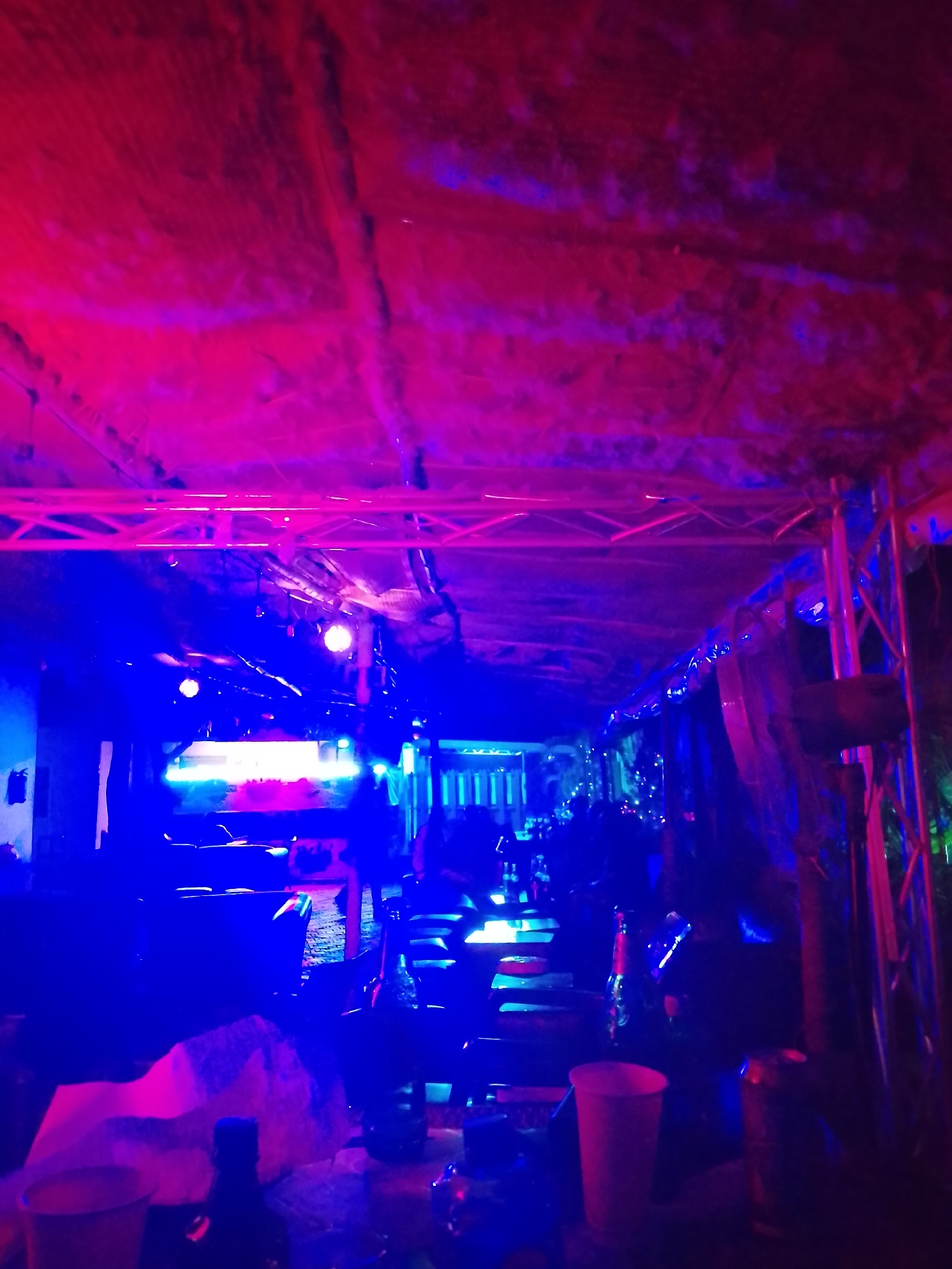 This image captures a vibrant night scene at an event venue in Yelwa, Nigeria. The space is bathed in intense artificial lighting, dominated by vivid magenta/red and electric blue hues, creating a dynamic and energetic atmosphere.
Above, a textured ceiling, possibly a tent or tarp, shows intricate patterns and supporting structures illuminated by the colored lights. Metal trussing, typical of stage or event setups, spans across the upper part of the frame, likely supporting various equipment.
In the midground, bright light sources, possibly stage lights or screens, emit a strong glow, further enhancing the dramatic lighting. Indistinct shapes suggest the presence of furniture or components of a performance area, such as a DJ booth or stage. People are visible as silhouettes in the background and midground, indicating a gathering or crowd immersed in the event.
The foreground features several objects on a surface, including disposable cups (one red, others translucent) and a bottle and can, suggesting active participation and beverage consumption at a social gathering.
The overall impression is that of a lively nighttime event, such as a concert, club night, or party, characterized by its striking light display and social ambiance. No discernible text is visible in the image.