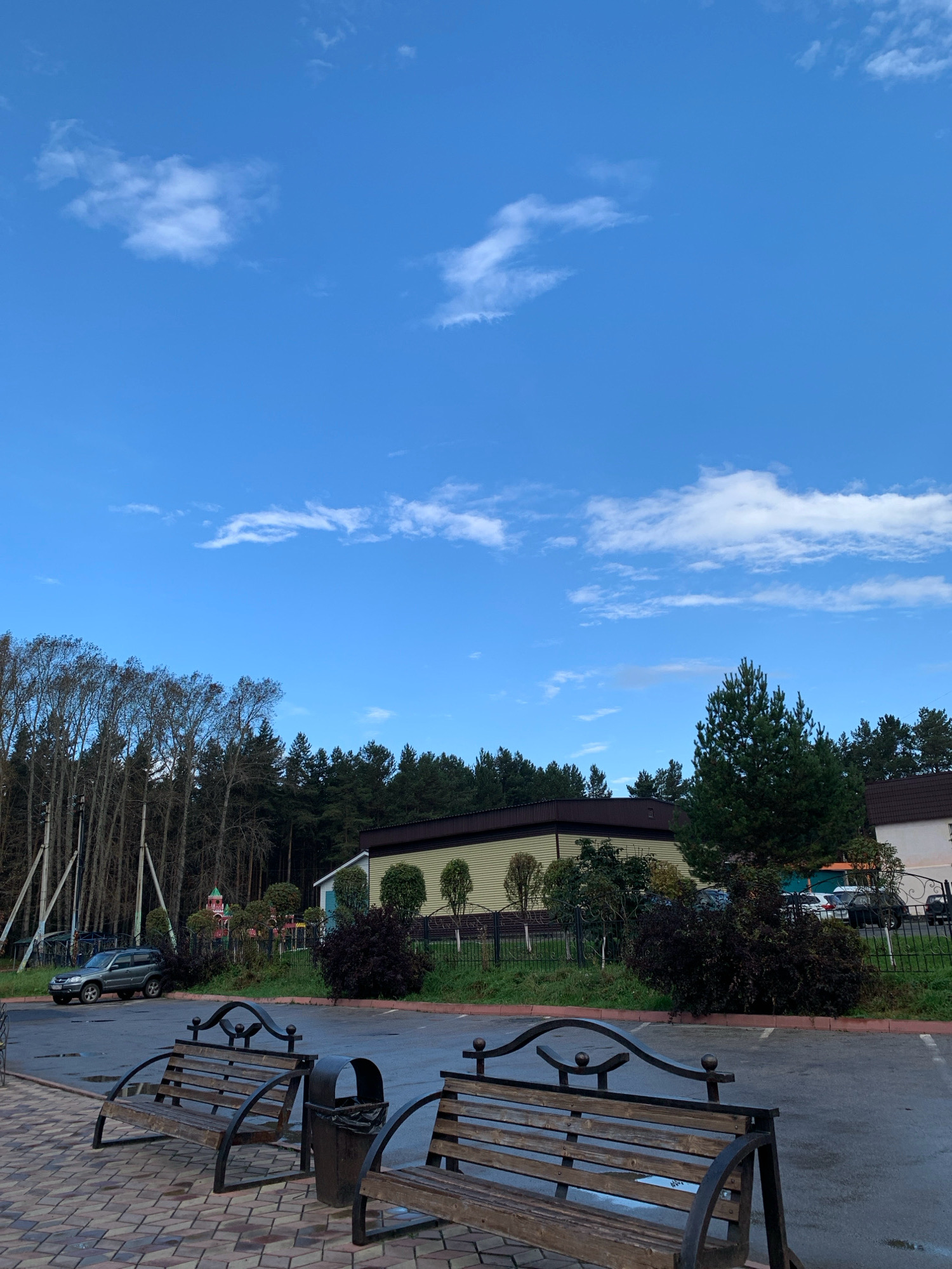 The image depicts an outdoor scene featuring two wooden benches with decorative black metal frames, positioned on a brick-paved area. Adjacent to the benches is a dark metal trash receptacle. Beyond this foreground, a paved parking lot is visible, with several parked vehicles, including a silver SUV. In the background, a line of trees, some bare and some evergreen, forms a dense forest. Further back, low-rise buildings with dark roofs are situated amongst the trees. The sky above is a clear blue with scattered white clouds. The environment suggests a public outdoor space, possibly a park or a recreational area, near a wooded region. There are no people or animals visible in the image. This scene is not specifically identifiable as being in Prokopyevsk, Russia.