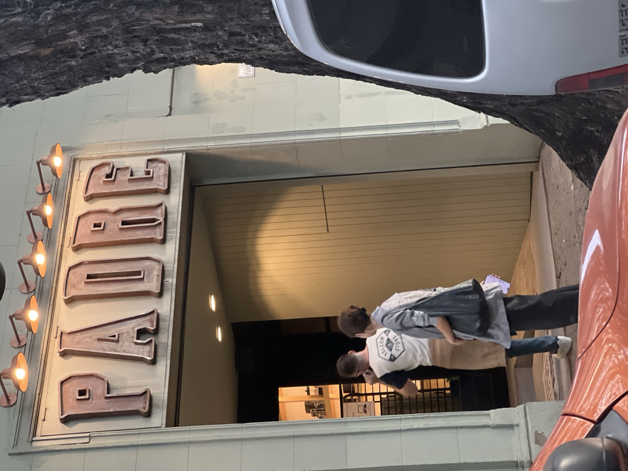 The image shows the entrance to a restaurant called "Padre" in Buenos Aires, Argentina. The sign "PADRE" is in large, lit-up letters on the left side of the entrance. Two people are standing near the entrance, one wearing a white t-shirt and the other wearing a gray jacket. A white car is parked on the left side of the frame, partially obscured by a tree. An orange car is on the right. The entrance leads into a dimly lit interior.