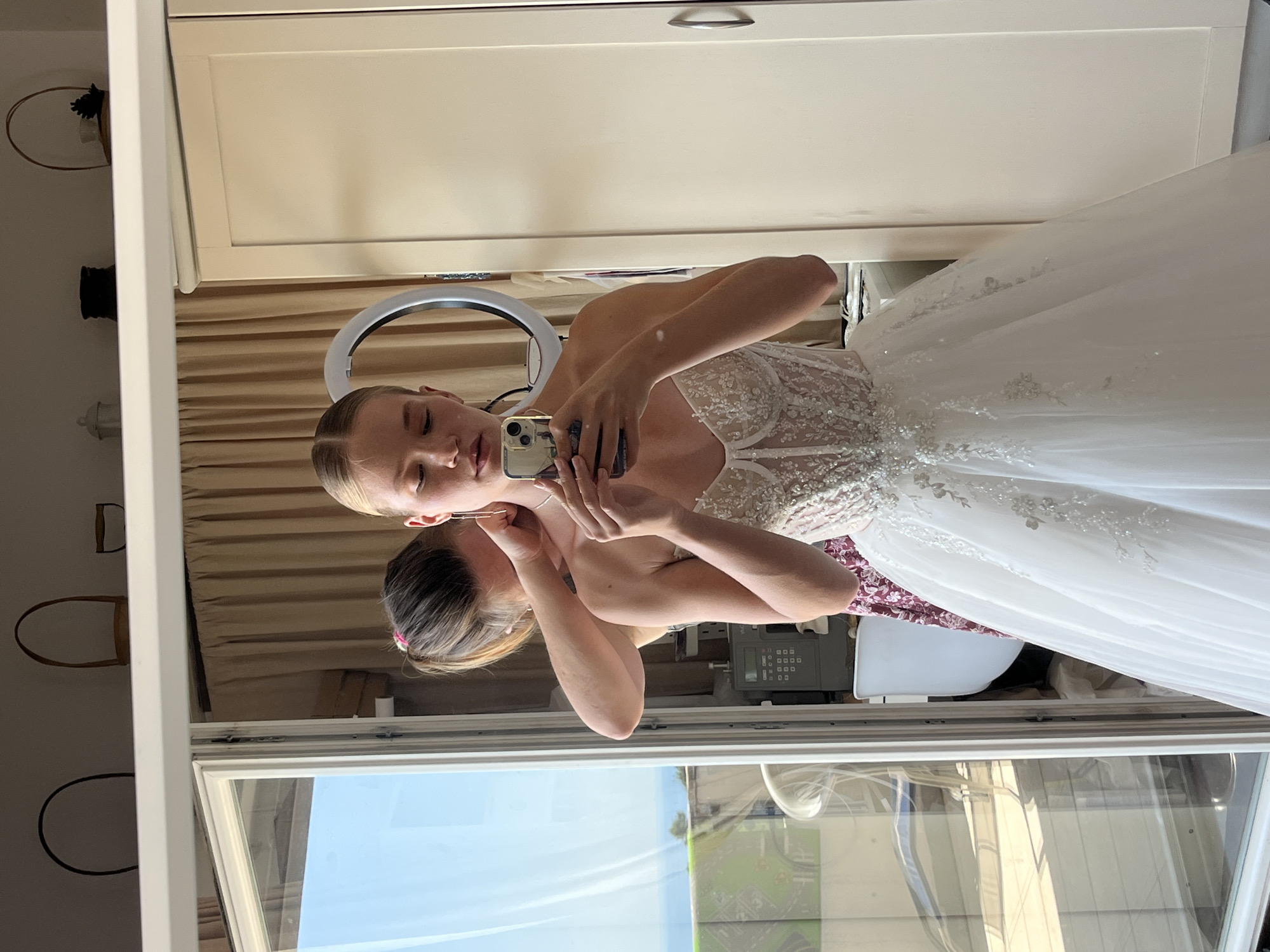This media file captures a young woman in a white wedding dress, taking a mirror selfie in what appears to be a bridal shop or a room set up for a wedding fitting. The woman has her hair pulled back and is wearing delicate earrings. She is holding her phone in her right hand, aiming it towards the mirror to capture her reflection.
The setting includes a white wardrobe or cabinet in the background. A ring light is visible, likely used for better lighting during photography. The dress itself is a formal white gown, with intricate beadwork and lace detailing on the bodice and a flowing skirt. A hint of a floral patterned garment is visible beneath the wedding dress.
The reflection in the mirror shows more of the room, including what looks like a window with natural light streaming in, suggesting it is daytime. The view outside the window hints at an outdoor scene, possibly with greenery and structures, which, given the location context of Budva, Montenegro, could be indicative of the local environment. There is no visible text in the image. The woman's expression is focused as she takes the photo, perhaps a mix of excitement and contemplation often associated with trying on a wedding dress.