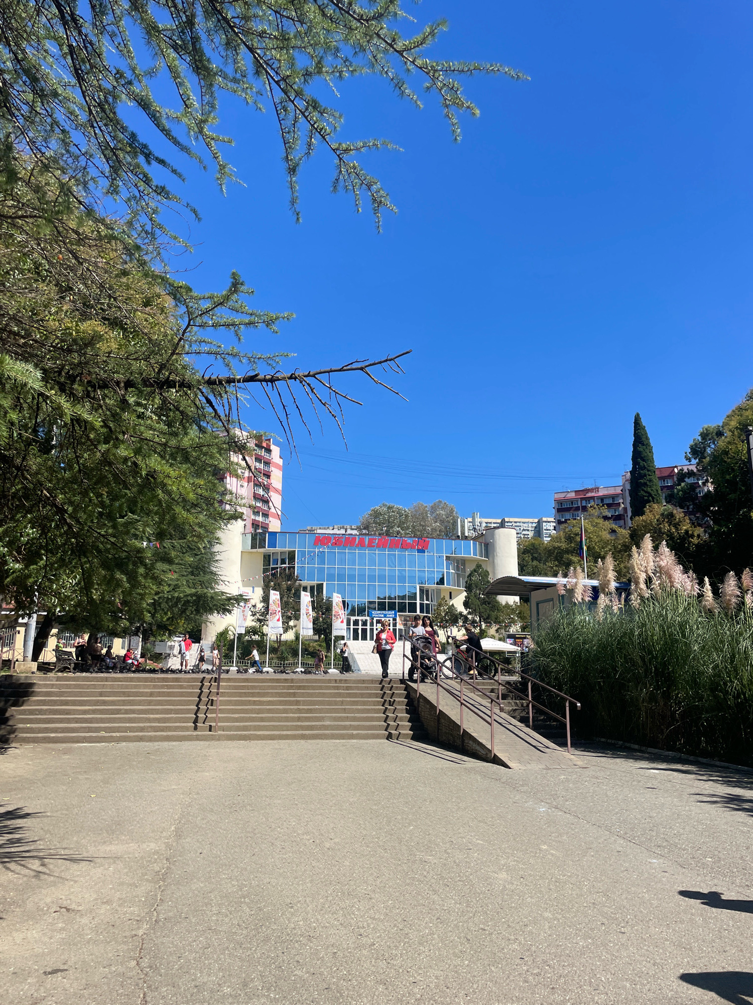 The image depicts a wide, sunlit plaza in front of a modern building with a blue glass facade and red lettering. A grand set of concrete stairs with handrails leads up to the building's entrance, and a sloping ramp is situated to the right of the stairs, also with handrails. Several people are visible on the stairs and in the plaza, some walking, others sitting or interacting with bicycles and a stroller. Tall green trees with dense foliage frame the left side of the scene, and a cluster of tall, feathery plants grows to the right of the ramp. In the background, multi-story buildings and a tall, slender cypress tree are visible against a clear blue sky. The overall atmosphere suggests a public space on a bright, clear day. The red lettering on the building appears to spell "РОССИЯ," which translates to "RUSSIA." This suggests the location is likely in Russia, and considering the architecture and vegetation, it is consistent with a resort area like Sochi.