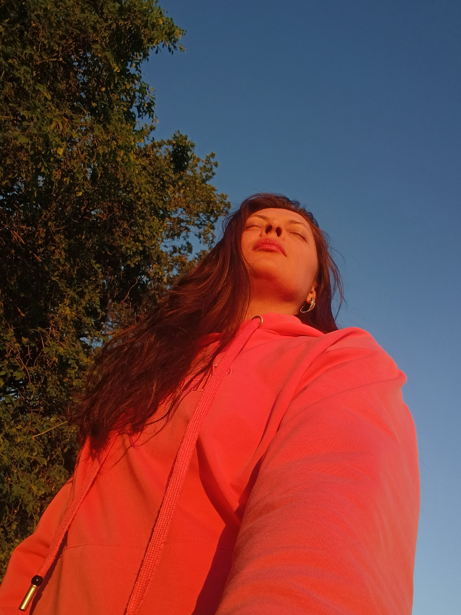 A young woman is depicted outdoors, with her head tilted back and eyes closed, illuminated by the warm glow of what appears to be sunset. She is wearing a bright coral-colored hooded sweatshirt with visible drawstrings. To her left, a large, leafy tree branches out against a clear blue sky. The low angle of the sun casts a warm orange hue across the woman's face and clothing. There are no discernible landmarks or indicators that place this image specifically within Saratov, Russia.