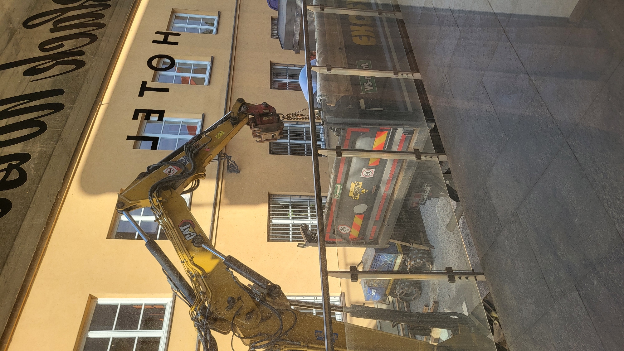 The image depicts a construction site with an excavator loading a dump truck. The excavator, a yellow CAT model, is positioned to the left, its arm extended over the dump truck. The dump truck is dark gray and has various markings, including "90 km" stickers and text that reads "VS-mont" and "EXOK". A person wearing a light blue shirt is visible inside the dump truck, appearing to be working. In the background, a yellow building with several windows can be seen. Black lettering, spelling "HOTEL", is affixed to the building above some of the windows. The scene is viewed through a glass railing, with dark gray tiled flooring in the foreground. The setting appears to be an urban environment, but the specific location in the city of Prešov, Slovakia cannot be determined from the visual information provided.