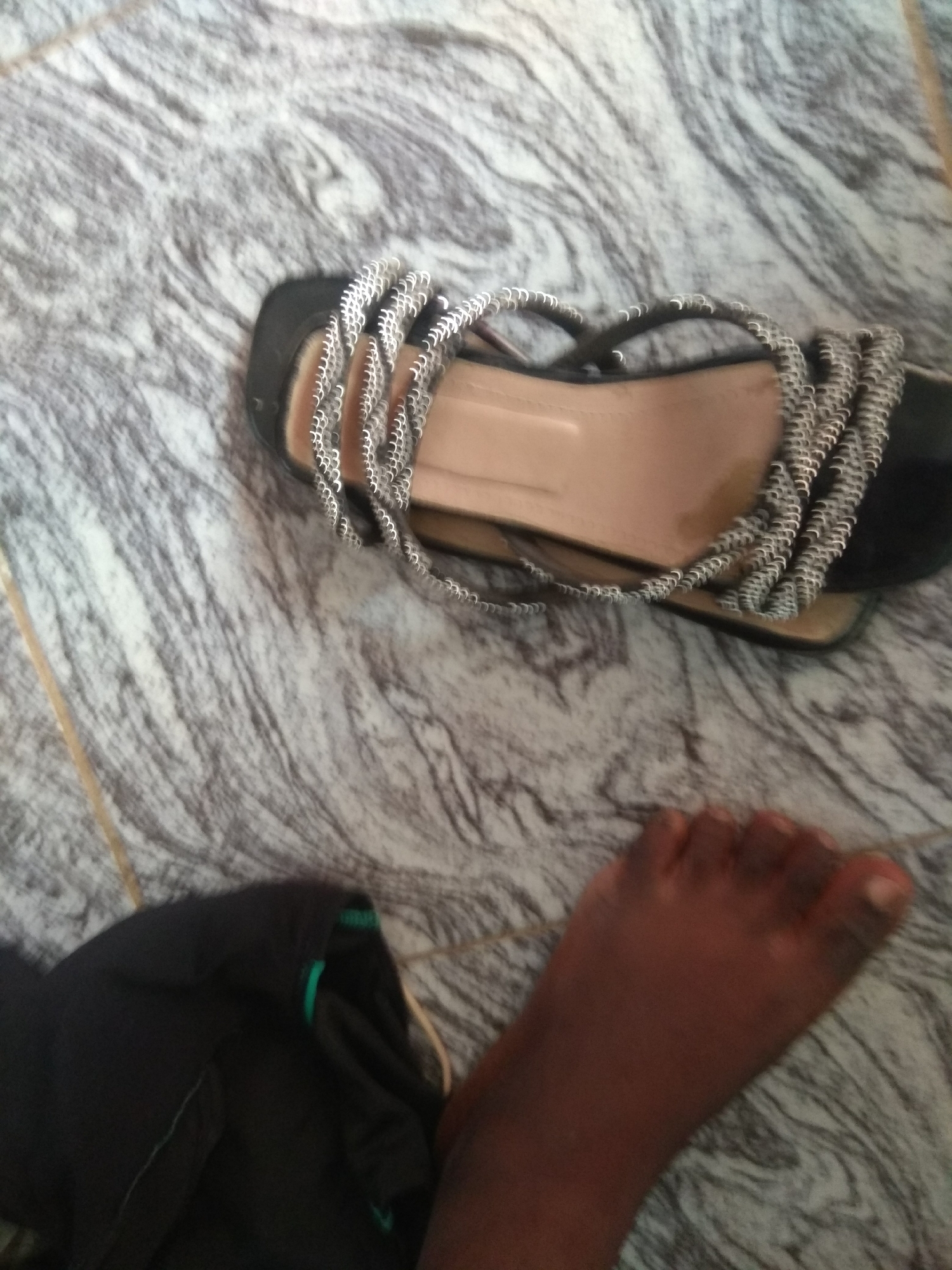 The image shows a close-up view of a person's foot next to a sandal. The foot is dark-skinned and appears to be positioned on a marble-patterned floor. A black sandal with silver decorative straps is lying on the floor near the foot. A piece of dark clothing is visible in the lower left corner of the image. The background is a floor with a grey and white marble pattern. The scene is likely indoors, possibly in a home or a retail space in Jalingo, Nigeria, given the prompt's context.