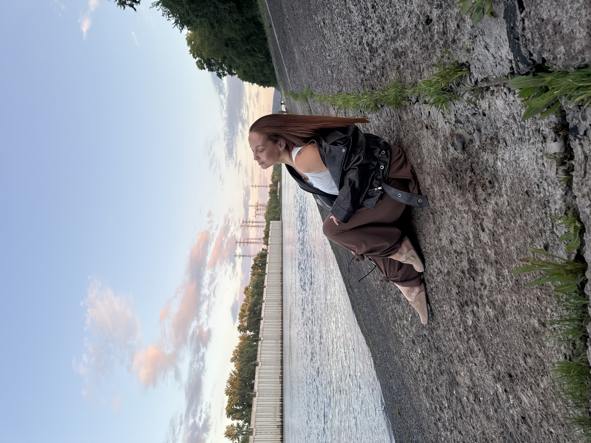 A young woman with long, reddish-brown hair is sitting on a rocky embankment next to a wide river. She is wearing a white tank top, a black leather jacket, and loose brown pants. Her head is tilted back, eyes closed, and her face is illuminated by soft, natural light, suggesting a moment of relaxation or contemplation. She is barefoot, with a pair of shoes resting near her feet. The river stretches to the left of the frame, with a long, concrete retaining wall visible on the opposite bank, lined with trees. In the distance, power lines and towers rise against a pale blue sky dotted with wispy pink and white clouds. The embankment is rough and textured, with cracks and patches of grass growing through. The scene appears to be set in the city of Chaikovsky, Russia, likely along the Kama River, given the prominent retaining wall and industrial power infrastructure.