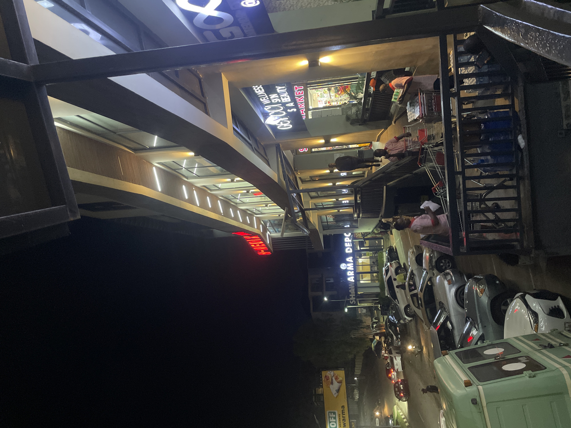 This image captures a nighttime scene outside a commercial building in Guzape, Nigeria. The building features a modern facade with illuminated signage. One prominent sign displays "GENOCY YELL" and mentions "WELLNESS," "SKIN," and "S A BEAUTY," suggesting a beauty or wellness establishment. Another sign reads "SHAWARMA DEPC," indicating a food vendor. The image shows a row of parked cars, including several silver sedans and a light green van, lining the street. People are visible in and around the building, some appearing to be customers or staff. One person in a pink shirt is seen near a shopping cart, and others are engaged in various activities within the illuminated interior spaces of the building. The overall atmosphere is one of a busy commercial area at night, with artificial lighting illuminating the scene against the dark sky. Visible text includes "H-MEDIX," "OFF," "akeaway," and "warma" on a billboard.