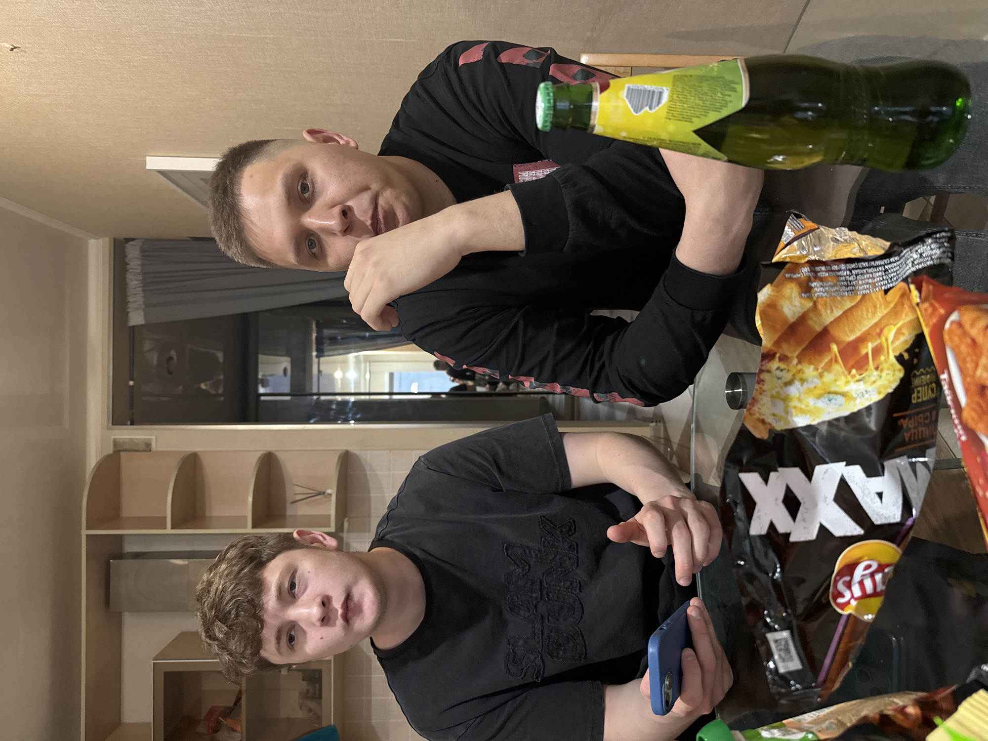 Two young men are sitting at a glass-topped table. One man is on the left and is holding a blue cell phone. The other man is on the right and his arm is raised to his head. The table is cluttered with open bags of Lay's Maxx chips, a green bottle of what is likely a beverage, and other partially-visible items. Behind the men, there is a room with cream-colored shelving and cabinetry. There is no indication of the location in Kazan, Russia.