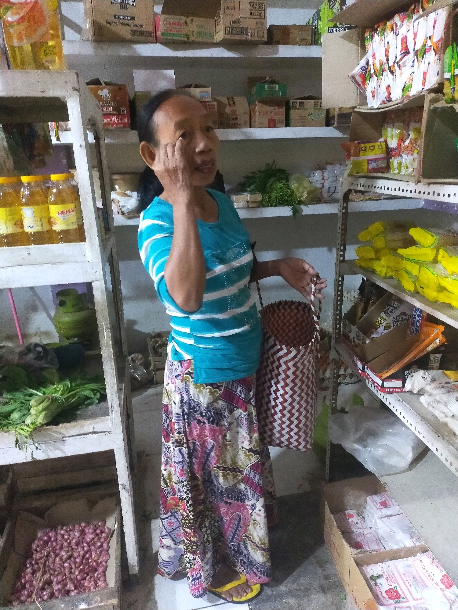 A medium shot captures a woman standing in what appears to be a small convenience store or warung in Mojodanu, Indonesia. She is positioned to the left of center, facing slightly to the right, and is holding a woven basket. The woman has dark hair and appears to be of middle age, with her right hand touching her face near her eye. She is wearing a teal t-shirt with horizontal white stripes and a long, colorful patterned skirt. Her feet are clad in yellow flip-flops.
The environment is densely packed with goods, suggesting a retail setting. To her left, a tall, open shelving unit displays several bottles of cooking oil. Below this, on a lower shelf, are green vegetables and a green gas cylinder. Further down, a crate is filled with red onions. To the woman's right, a metal shelving unit is stocked with various products. The top shelf has an assortment of yellow-wrapped items. The shelves below contain boxes of packaged goods, including what appear to be instant noodles and bags of flour. The floor is concrete. The lighting is dim, with artificial light sources illuminating the scene. The overall impression is that of a modest, well-stocked local shop.