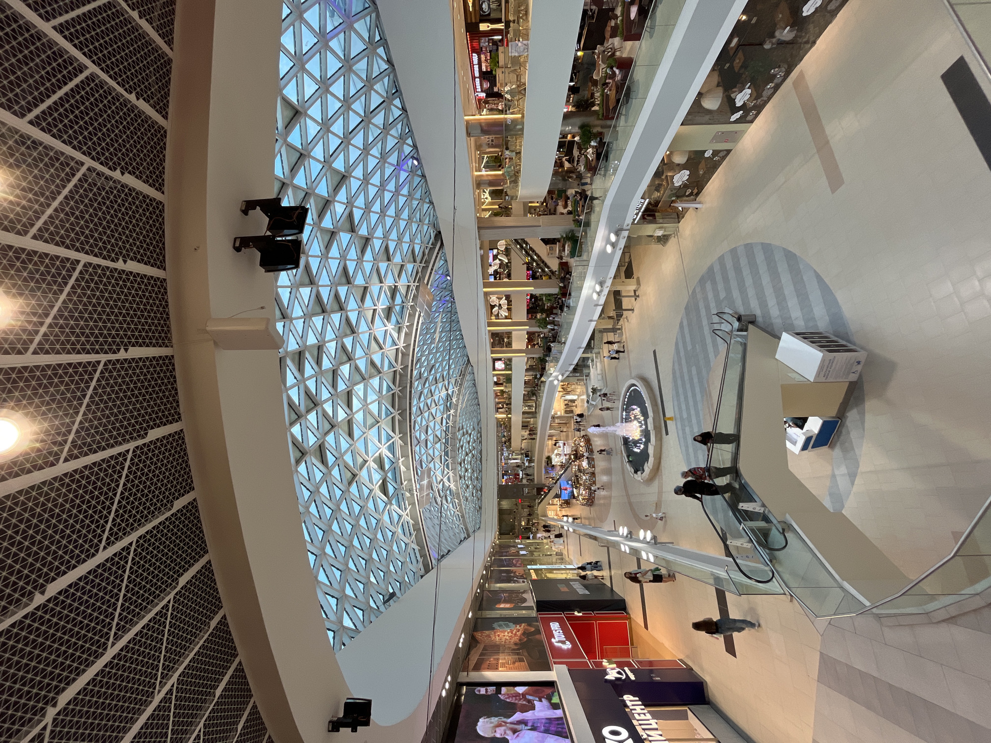 An interior view of a large, modern shopping mall in Sochi, Russia, is depicted. The mall features a multi-level design with escalators and walkways connecting different sections. A prominent glass ceiling with a geometric pattern allows natural light to illuminate the spacious interior. In the center of the ground floor, a fountain with cascading water is visible, surrounded by shops and seating areas. People are scattered throughout the mall, walking, shopping, and sitting at tables. Several storefronts are identifiable, including one with a large digital display showing a person. The overall atmosphere is one of a bustling commercial hub.