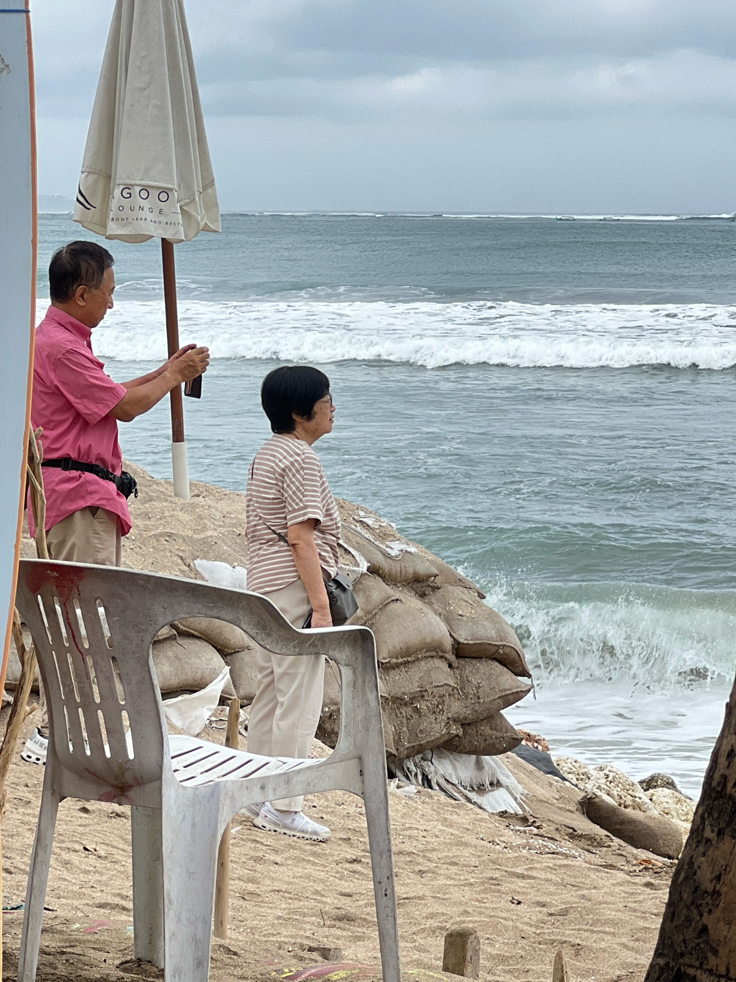 An overcast day at Kuta Beach, Bali, Indonesia. A man in a pink shirt and khaki pants stands to the left, holding a phone in his right hand as if taking a photograph. Beside him, a woman in a striped shirt and khaki pants looks out at the ocean. They are positioned on a sandy embankment reinforced with sandbags. A white plastic chair sits in the foreground, facing away from the camera. In the background, waves crash onto the shore under a cloudy sky.