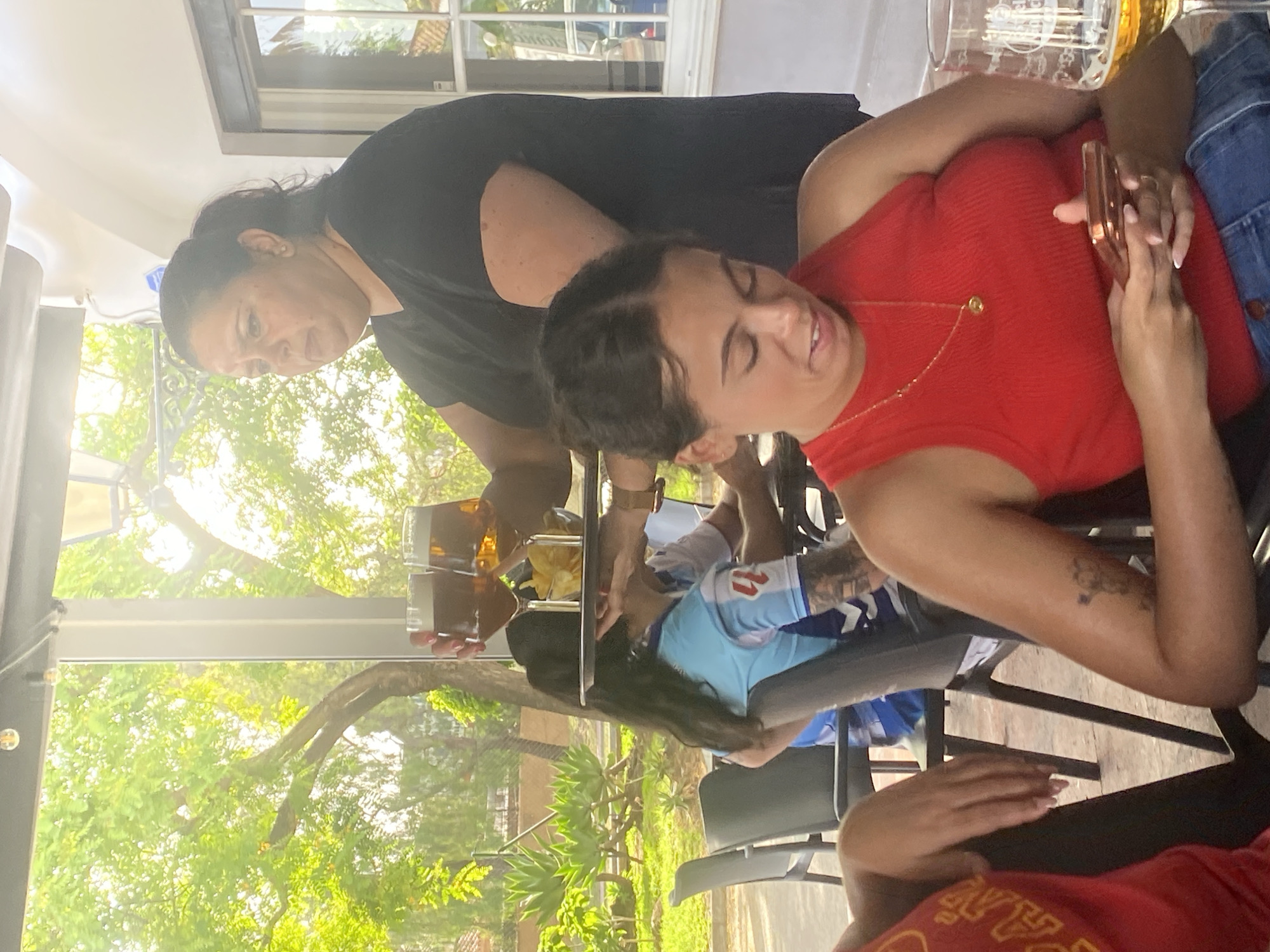 The image shows a group of people at an outdoor restaurant in San Pedro De Alcántara, Spain. A woman in a black t-shirt is serving drinks to patrons at a table. She is holding a tray with two glasses of beer and a bowl of chips. In the foreground, a woman in a red tank top is looking at her phone. Another person in a red shirt is partially visible on the left. The background shows lush greenery and other diners. The ambiance suggests a casual dining experience on a sunny day.