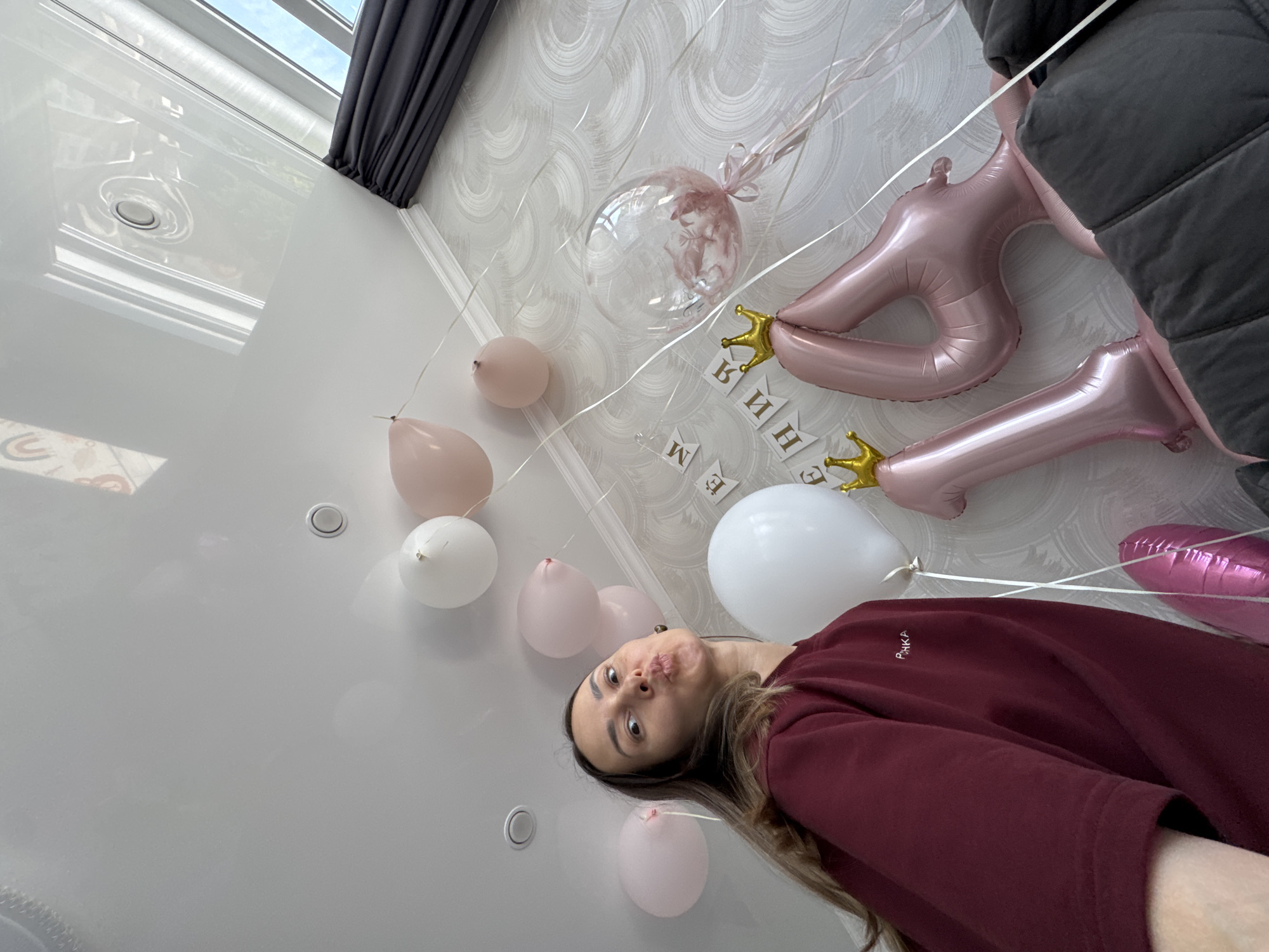 The image shows a woman taking a selfie in a room decorated with balloons for a celebration. The balloons include pink and white colors, with one clear balloon containing pink feathers. There is a pink balloon shaped like a number "1" with a gold crown decoration. A banner spelling out the name "Alina" in Russian is visible. The room has white walls and a white ceiling with recessed lighting. A gray curtain can be seen at the top of the frame. The woman is wearing a burgundy sweatshirt with Russian lettering. She is looking at the camera with a puckered expression. The location of the image is not possible to determine as Sochi, Russia, from the visual data in the image.