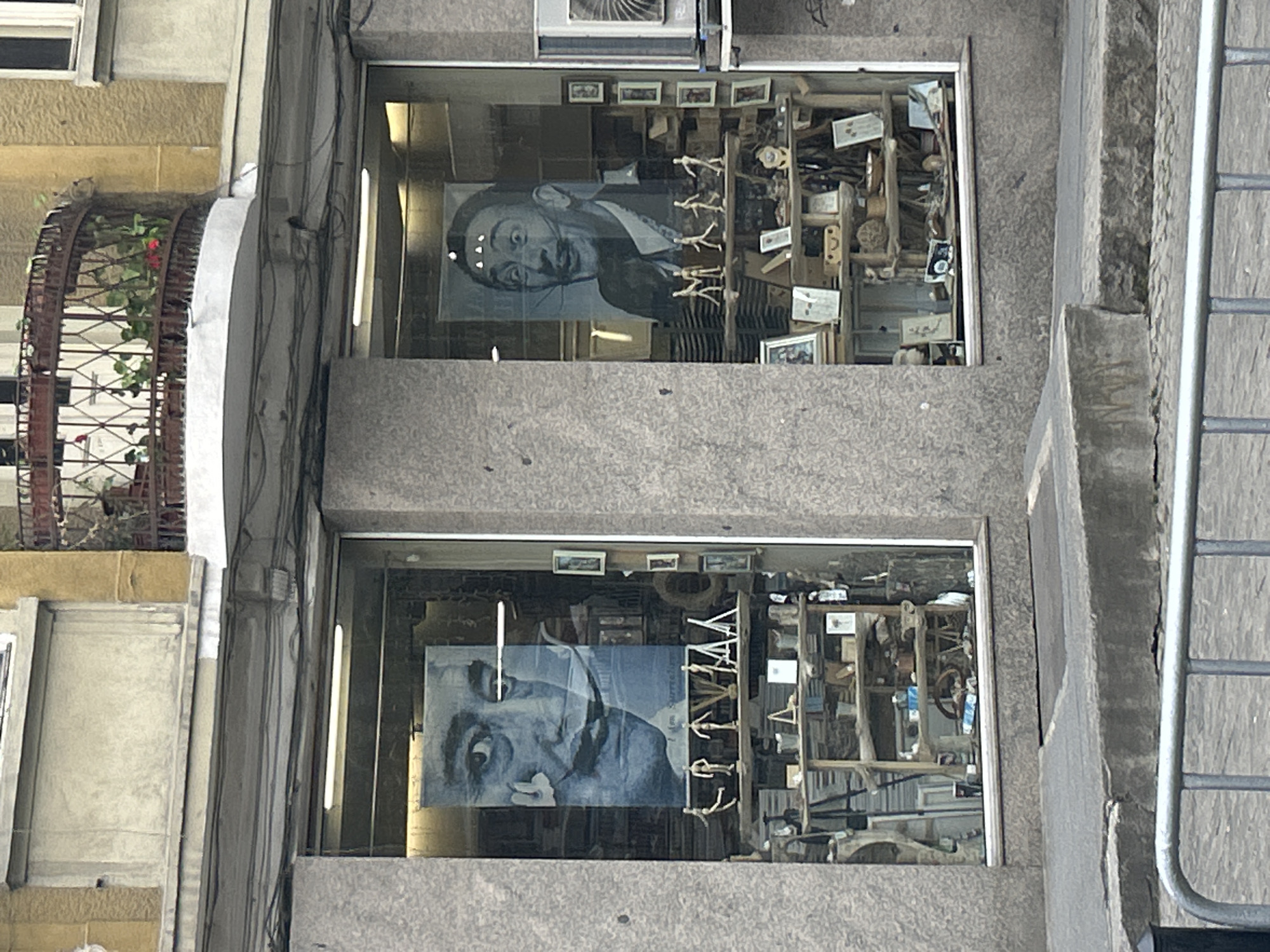 The image shows a shopfront in Plovdiv, Bulgaria, featuring two stacked display windows. Each window contains a portrait of Salvador Dali, one in black and white and the other in shades of blue. The windows also hold an assortment of artifacts, including small pictures and wooden models. The building exterior is concrete. A balcony with a decorative metal railing and flowering plants is visible to the left of the shopfront. Metal handrails are situated to the right of the shopfront.
