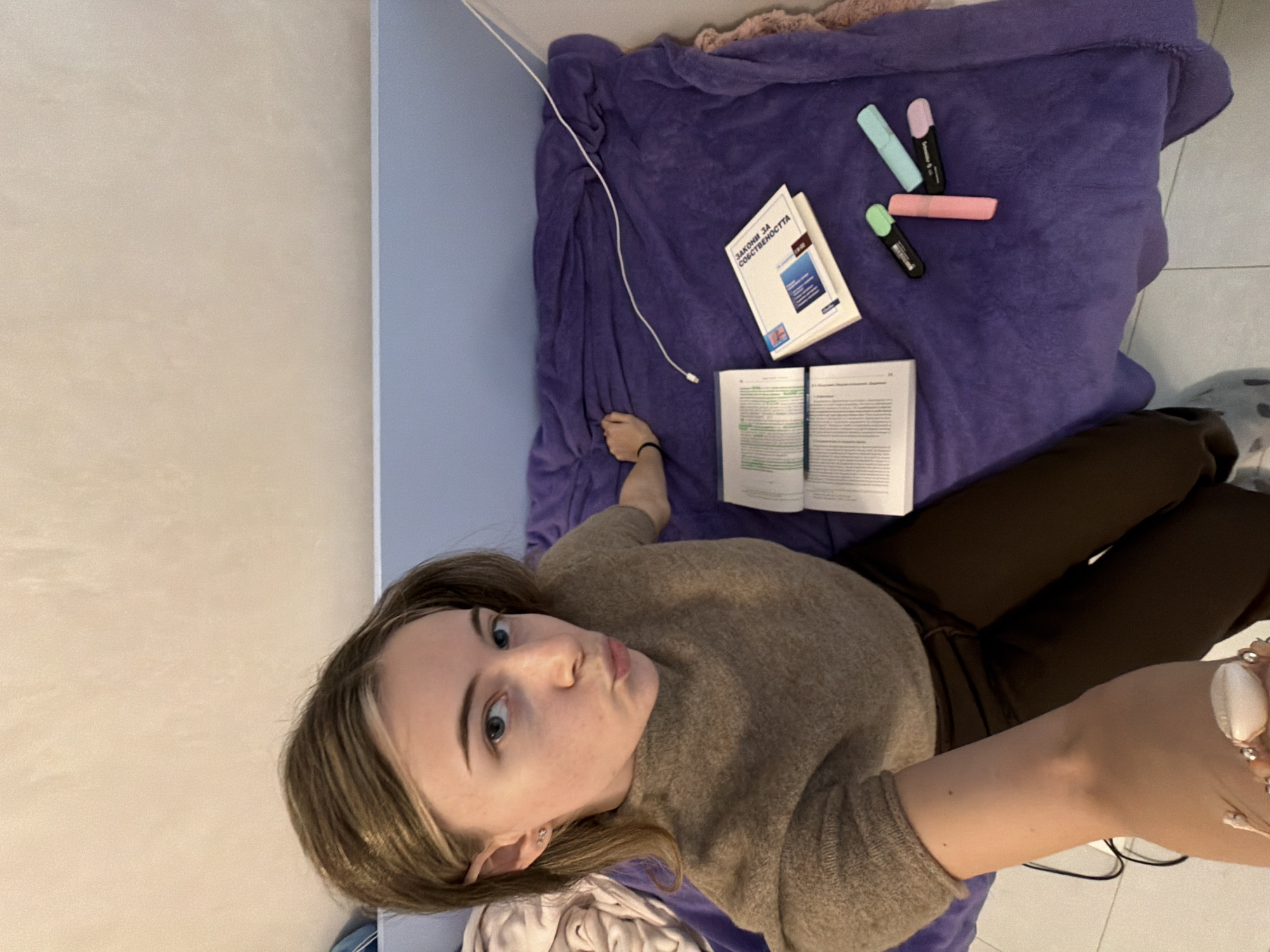 The image captures a top-down view of a young woman lying on what appears to be a bed or floor, surrounded by study materials. The setting is indoors, with light beige and light blue walls visible in the background, suggesting a room.
The central figure is a young woman with light brown hair and blue eyes. She is looking directly up at the camera with a casual, slightly puckered expression, as if posing playfully. She is wearing a light brown or beige long-sleeved knitted sweater and dark brown trousers. On her left wrist, a thin black bracelet is visible, and on her right wrist, she wears a more decorative bracelet featuring large white, shell-like beads interspersed with smaller silver-colored beads.
She is lying on a soft, plush purple blanket or covering. Next to her, several items are laid out:
- A closed book with a white cover, clearly displaying the title "ЗАКОН ЗА СОБСТВЕНОСТТА" (which translates to "Law of Property" in Bulgarian).
- An open textbook, with pages of text visible, suggesting it's actively being read or studied.
- Three highlighters (one light blue, one light pink, and one light green) are clustered near the closed book.
- A thin white cable, likely a charger for an electronic device, snakes across the purple blanket.
- A pair of black-framed eyeglasses are partially visible near her right arm.
The overall scene suggests a moment of relaxation or a break from studying. The lighting is even and bright, indicating an indoor environment, possibly during the day or well-lit by artificial lights.