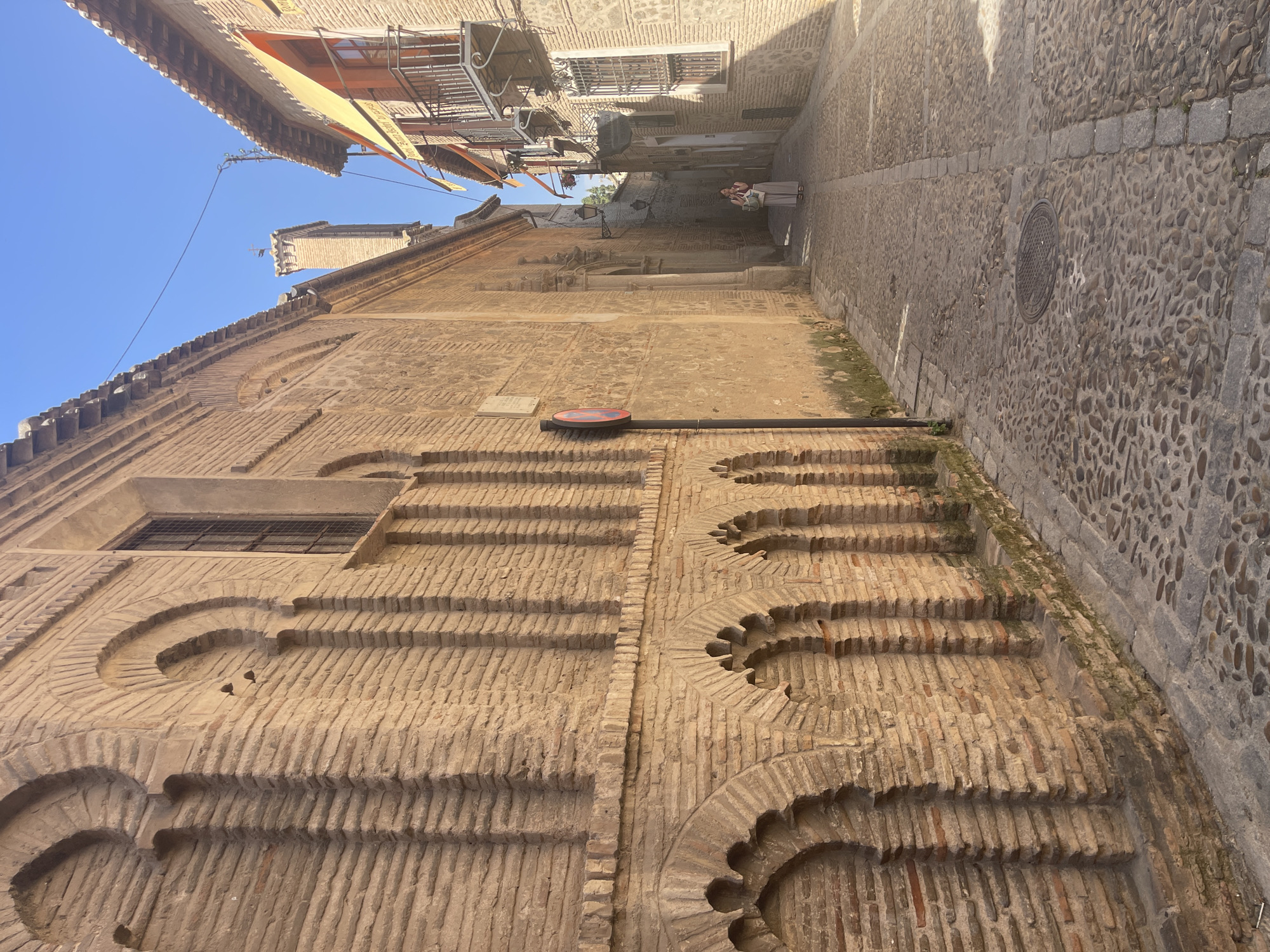 An eye-level, full shot shows a narrow, cobblestone street in Toledo, Spain. The street is flanked by tall, brick buildings with arched windows and a few balconies. The sun casts shadows on the street, indicating it is daytime. A woman in a light-colored outfit stands in the middle of the street, looking down. A red and white traffic sign is posted on the left side of the street. The architecture suggests a historical district, possibly with Moorish influences given the horseshoe arches visible on the building to the left. The sky is clear and blue.