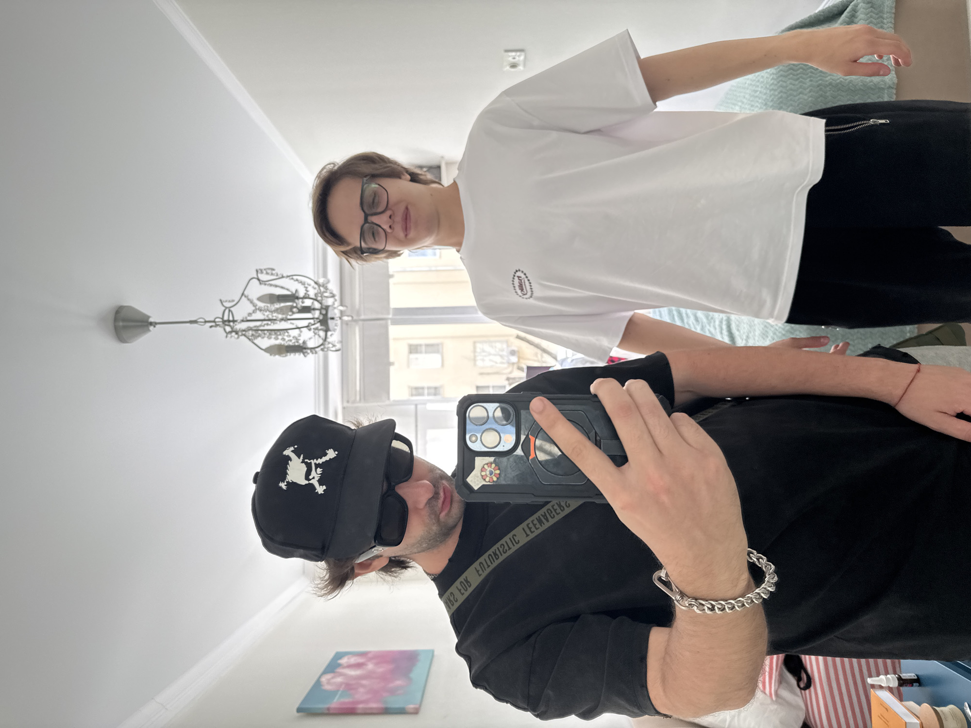 Two young men are posing for a mirror selfie in what appears to be an apartment. The man on the left is wearing a black t-shirt, a black baseball cap with a white skull design, and black sunglasses. He is holding a smartphone in his right hand, which is angled towards the mirror. He also has a silver bracelet on his left wrist. A black strap with white lettering is slung across his chest. The man on the right is wearing a white t-shirt with a small embroidered logo and black pants. He has on glasses and is smiling. The room is well-lit, with a chandelier hanging from the ceiling and a painting on the wall. The overall impression is casual and informal, possibly a moment captured between friends. The location is not identifiable as Moscow, Russia.