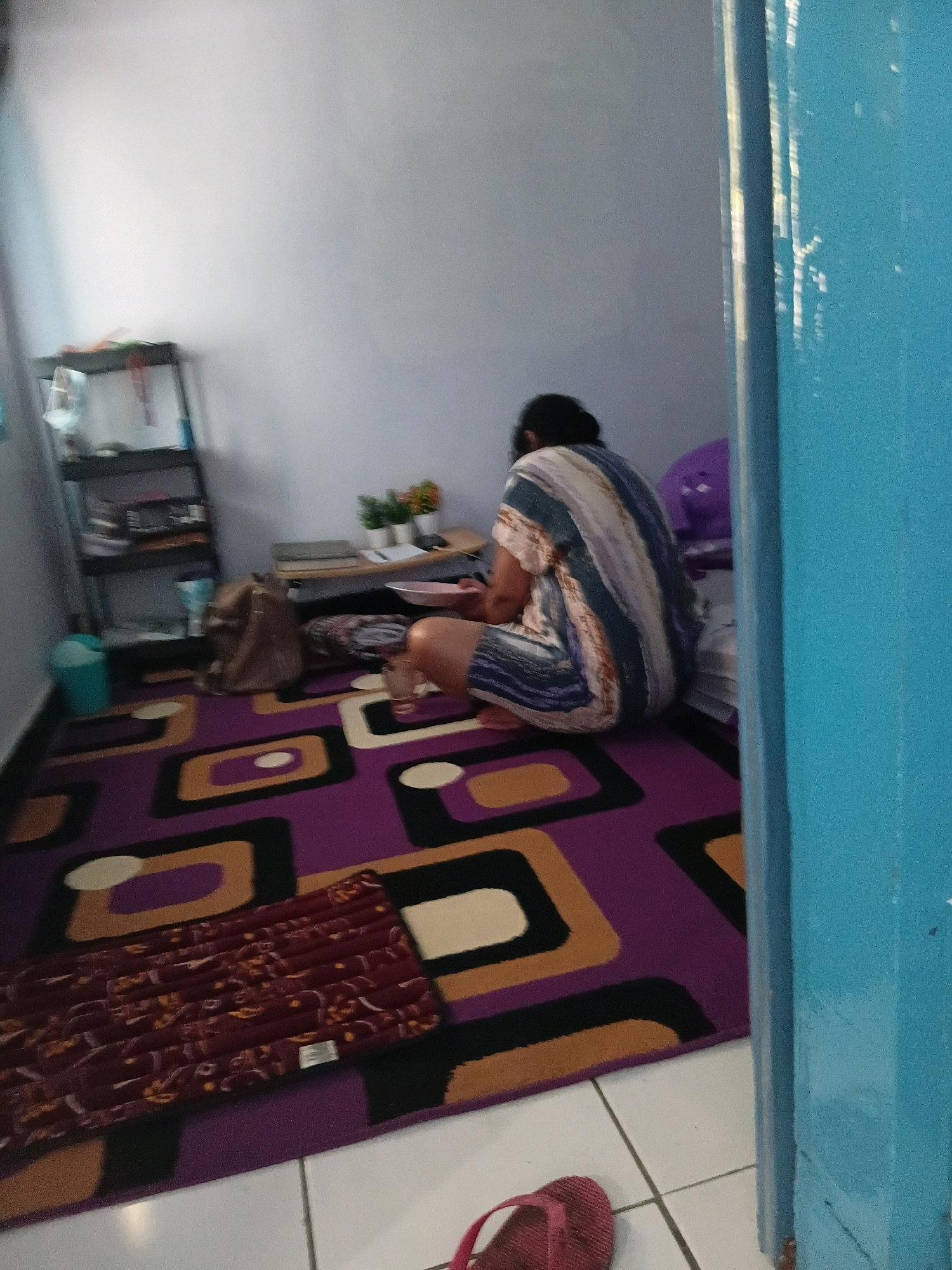 The image depicts an indoor scene, likely a bedroom or living area, with a focus on a person and their surroundings. The room features a light-colored wall and a vibrant purple rug with an abstract geometric pattern. To the left, a black shelving unit holds various items, including what appears to be books and a blue trash can. A brown bag is placed on the floor near the shelves. In the center, a person is seen from behind, wearing a patterned dress. They are squatting or kneeling on the rug, with their back to the camera, facing a low table or desk. On this surface, there are two potted plants, books, and a white bowl. To the right of the person, a white mattress or cushion is visible, and a portion of a blue door frame is present in the foreground, partially obscuring the scene. A red flip-flop is visible on the floor near the door. The lighting suggests an indoor environment during the day. The overall impression is a candid shot of someone engaged in an activity within a domestic setting. The mention of Tanggul Wetan, Indonesia, places this scene within a specific geographical context, suggesting it is a residential interior in that location.
