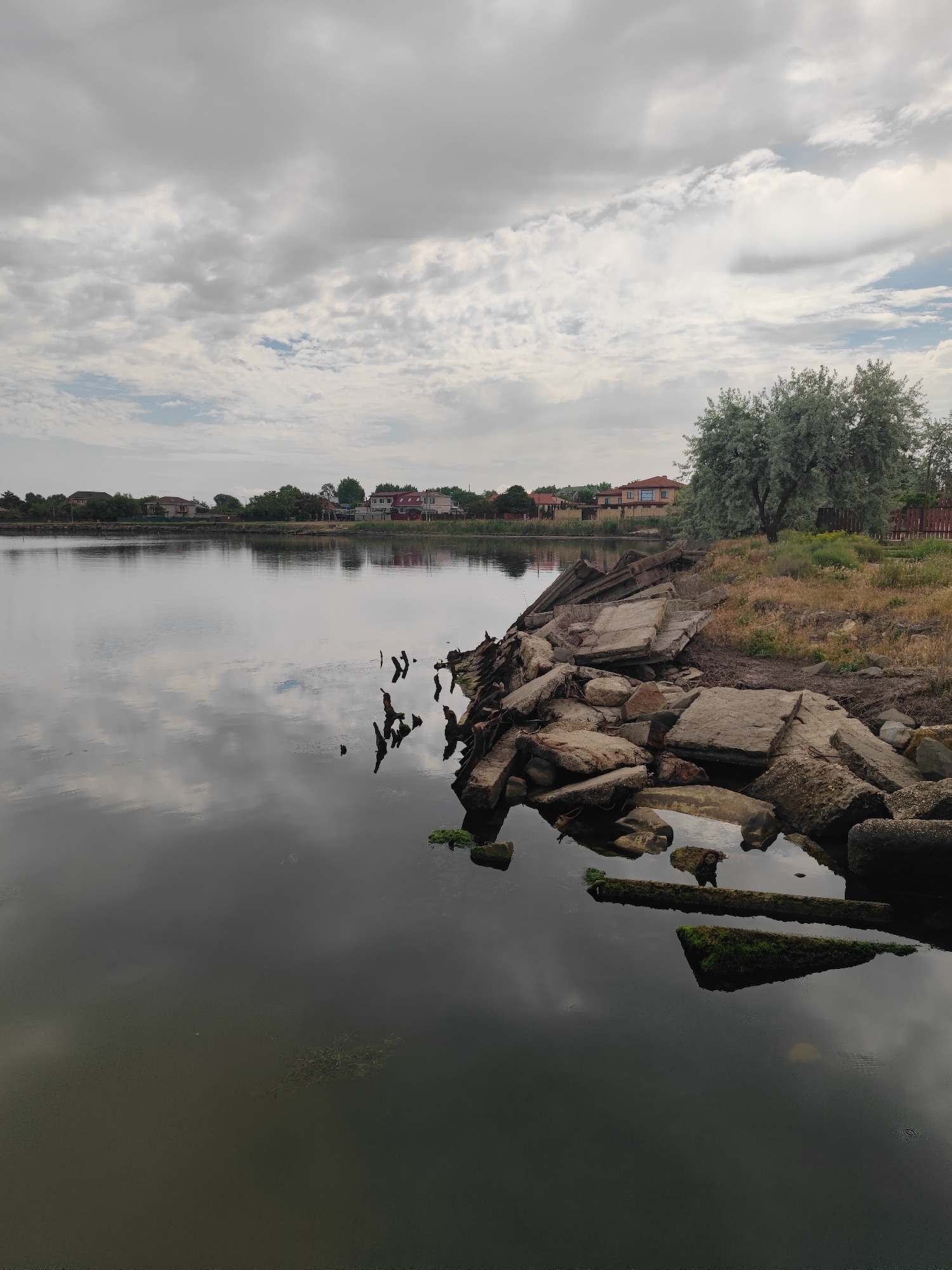 The image depicts a body of water, possibly a lake or a wide river, in Sennoy, Russia. The sky is overcast with grey clouds reflected on the water's surface, obscuring its depth. Along the right edge of the water, there's a shoreline with scattered, broken concrete slabs and rocks. Some of the concrete fragments are partially submerged and have patches of green algae. In the background, there is a line of houses, suggesting a residential area. A few trees are present on the shoreline and behind the houses. The overall scene has a somber or bleak atmosphere, likely due to the weather and the deteriorated state of the shoreline.