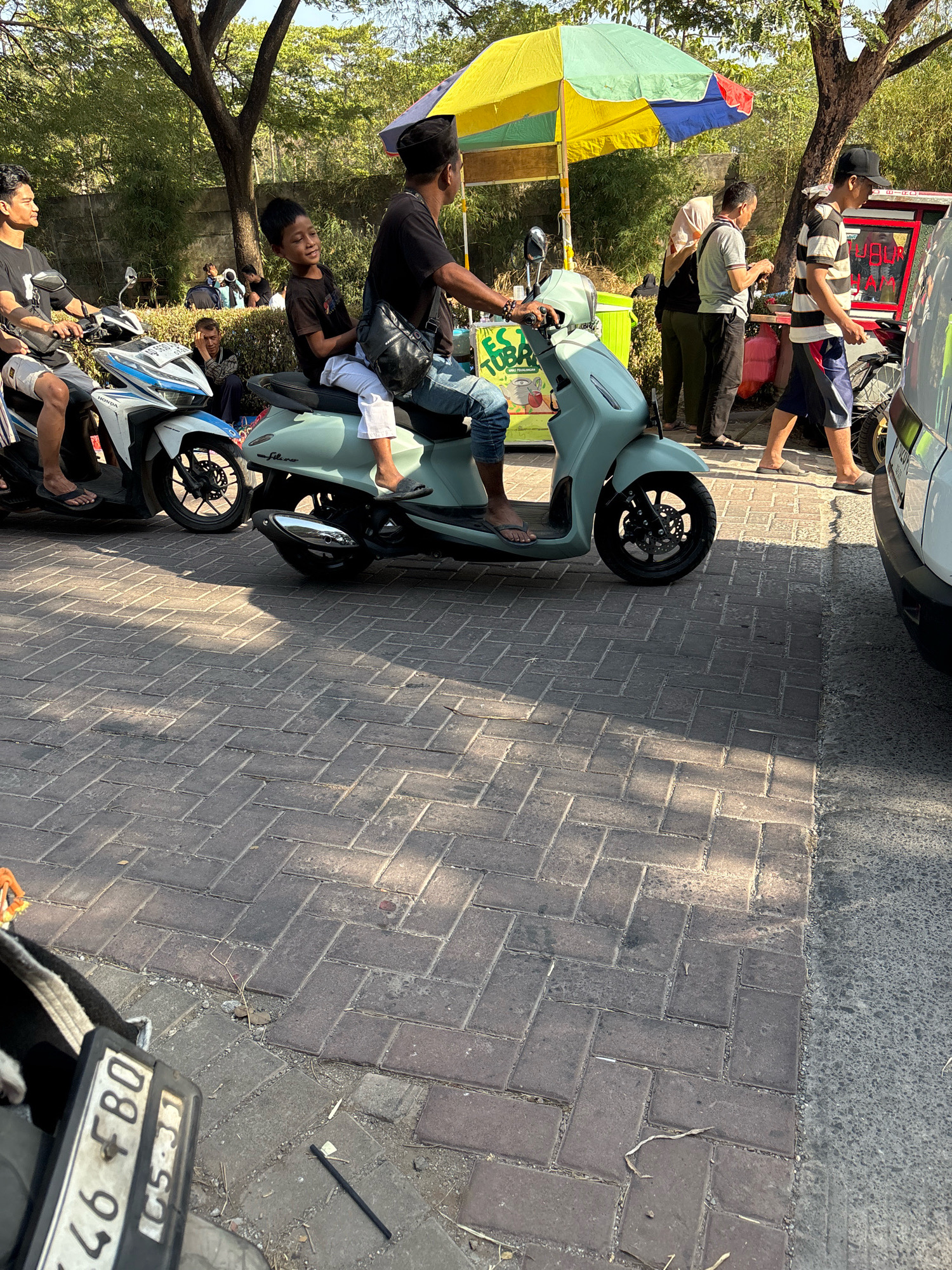 In Telagamurni, Indonesia, an outdoor scene unfolds on a brick-paved surface, bathed in sunlight creating distinct shadows. Two men are positioned on a light green scooter, with the adult driver wearing a black t-shirt and a black cap, and a younger boy in a dark t-shirt and white pants seated behind him. The boy is looking to his right with a slight smile. Adjacent to them, another man on a white and blue Honda scooter is partially visible, wearing a black t-shirt and light-colored shorts.
In the background, a colorful umbrella, primarily yellow, green, and blue, shades a food stall. Several other individuals are present, including a woman in a light pink headscarf and dark clothing, and men in casual attire. One man, wearing a striped shirt and shorts, walks towards the food stall. A white vehicle is parked on the right side of the frame, obscuring part of the scene. The environment is characterized by trees and foliage, suggesting a park-like setting or a roadside with greenery. A license plate, partially visible on a motorcycle in the foreground, reads "46 FBO C5331".