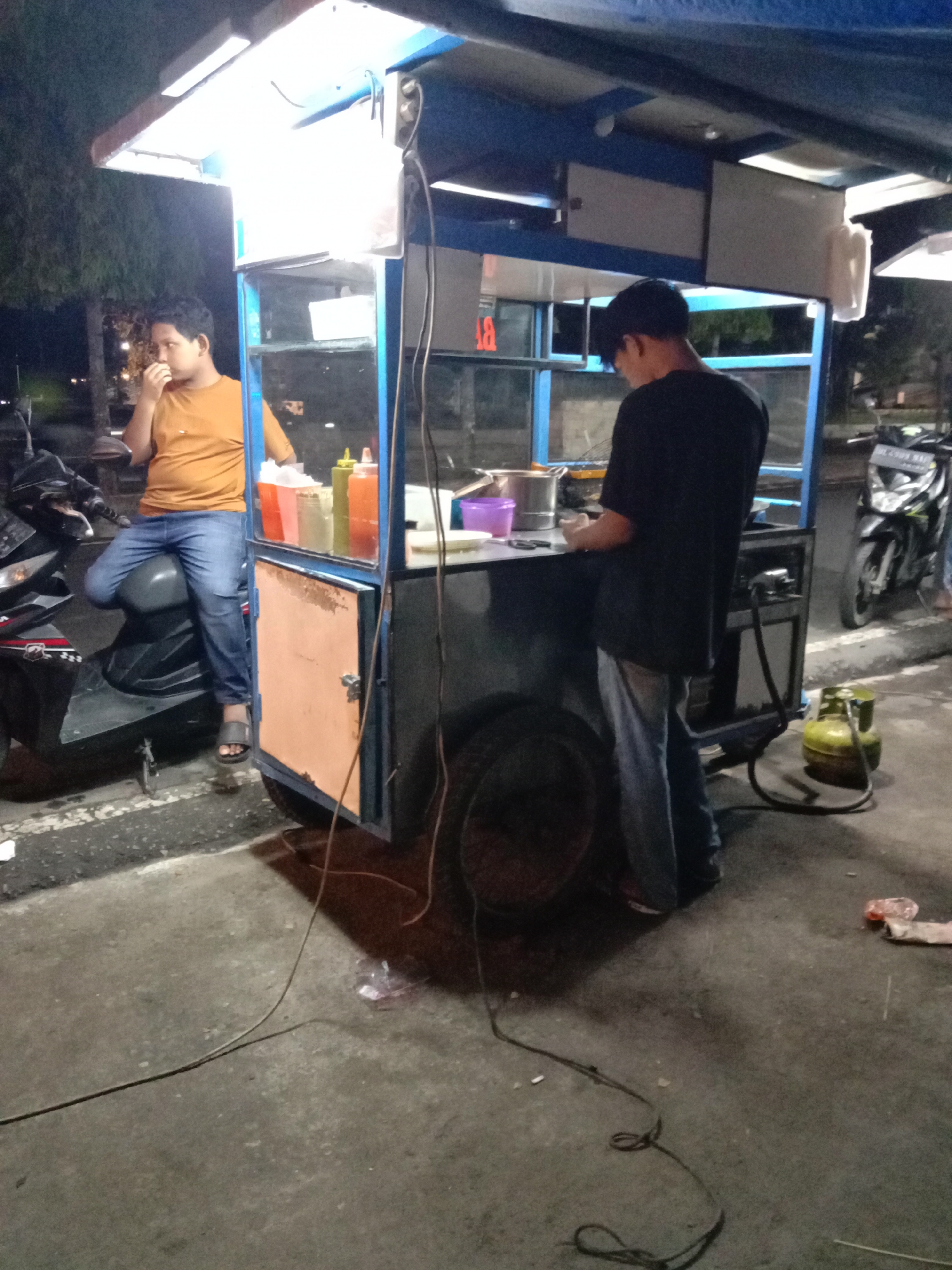 The image shows a food cart in what appears to be Lhokseumawe, Indonesia. The cart, illuminated by a light overhead, is being operated by a person wearing a black shirt and jeans who is presumably preparing food. Another person, wearing an orange shirt and jeans, is sitting on a parked black scooter next to the cart, seemingly waiting or watching. Condiments are visible on the cart. A green gas cylinder is positioned near the cart, presumably for cooking. Power cords are on the ground. Another scooter is visible in the background. The scene appears to be at night.