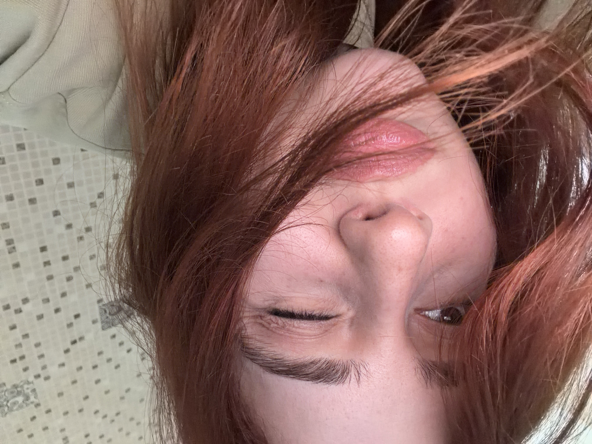 A close-up, upside-down image shows a person with fair skin and reddish-brown hair. The person's left eye is mostly closed, while the right eye is partially open, revealing a brown iris. The person has full lips with what appears to be lip gloss on them. The background shows a surface with a small, patterned tile. The city of Tbilisi, Georgia is not discernible from this image.