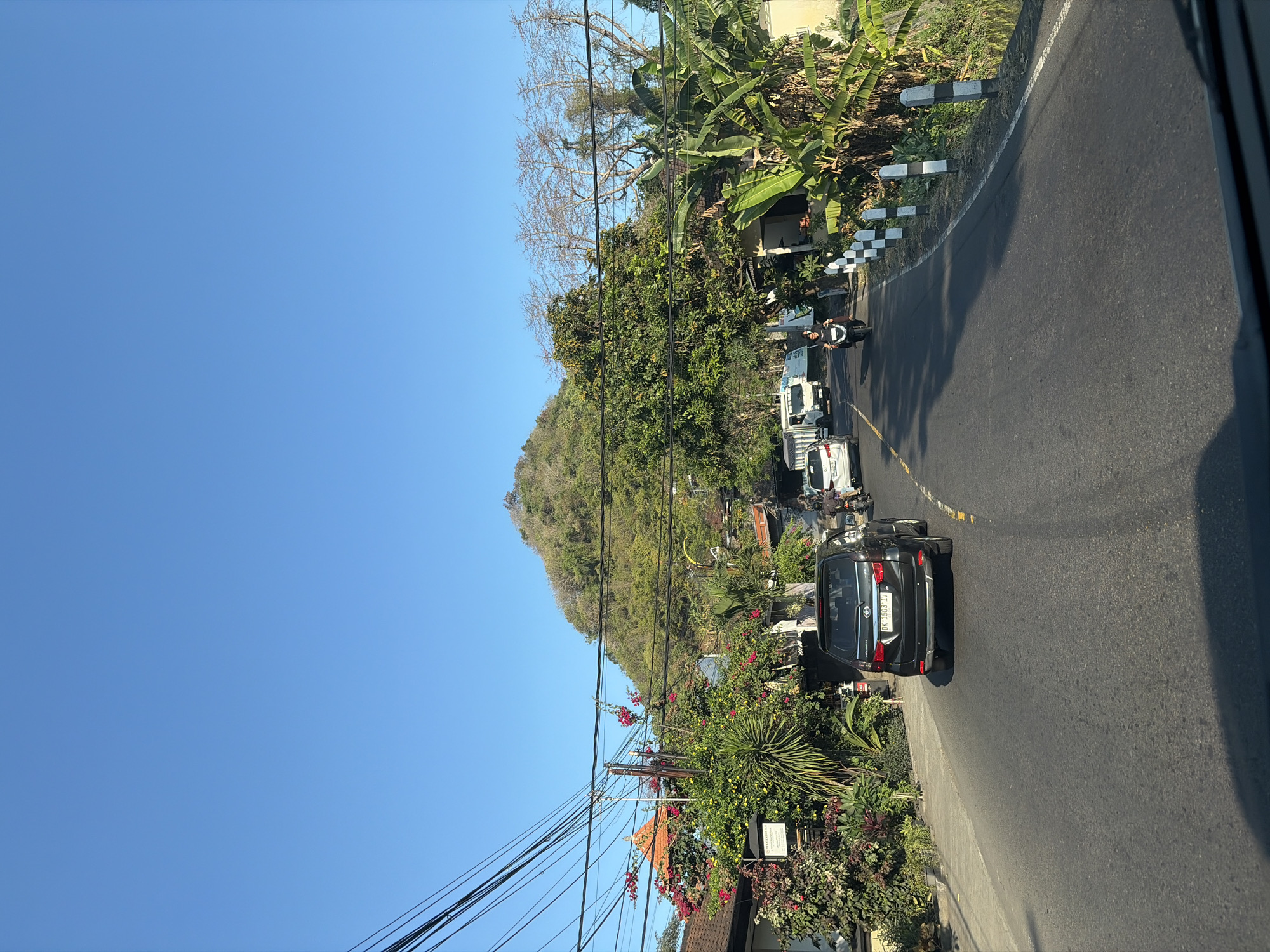 The image captures a roadside view in Karangasem, Indonesia, under a clear blue sky. A paved road curves gently to the right, with a dark SUV in the foreground. Further down the road, a white truck and a motorcycle with a rider are visible. On the left side of the road, a building with an orange-tiled roof is partially obscured by lush green foliage and vibrant pink and red flowers. Several utility poles with a tangle of black wires crisscross the upper portion of the image. On the right side, dense vegetation, including banana trees, lines the roadside, and a series of black and white striped bollards demarcate the edge of the road. In the background, a tree-covered hillside dominates the landscape, with a peak rising against the horizon.