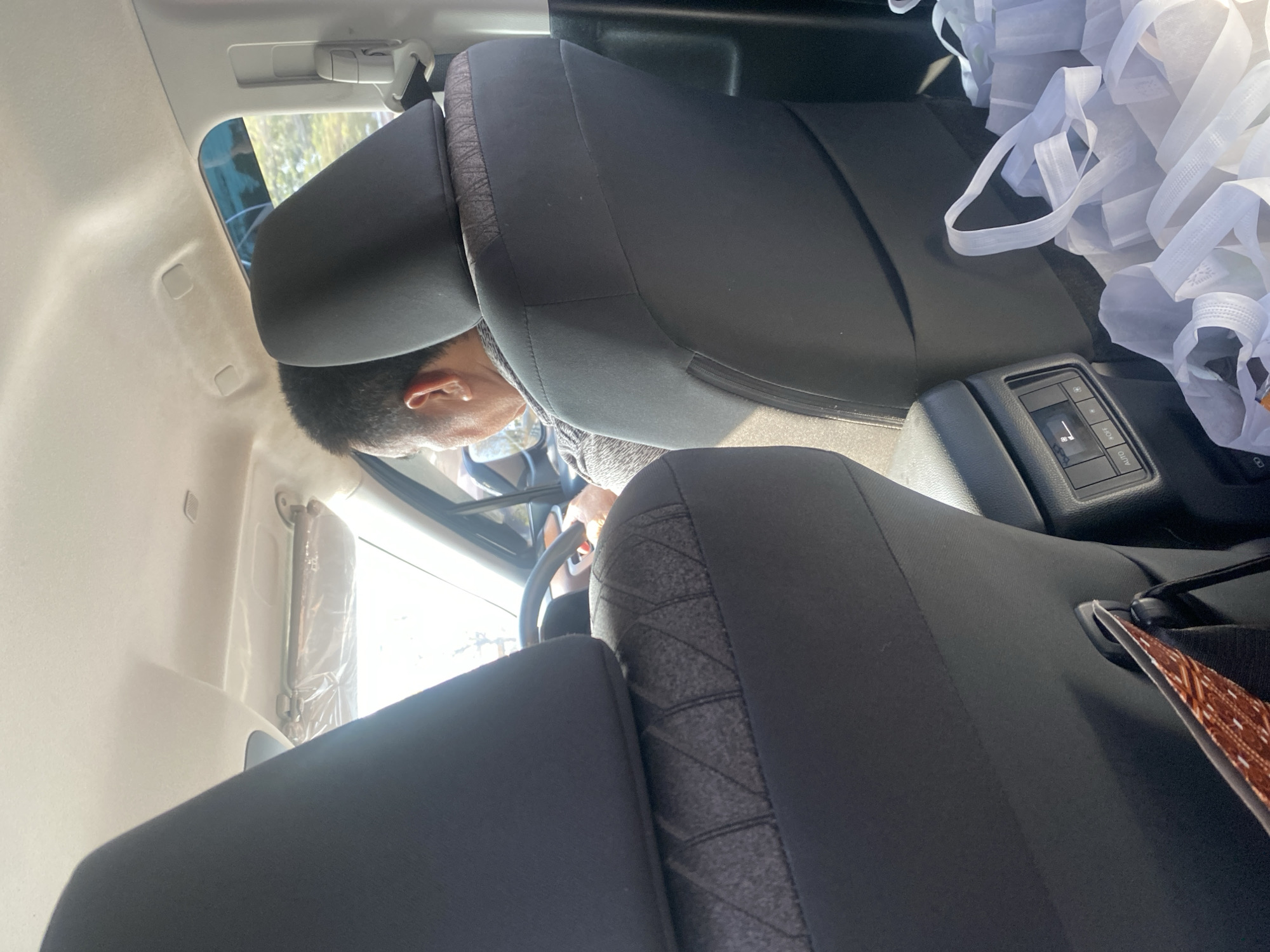 The image shows the interior of a car, viewed from the back seat. A man is visible in the driver's seat, seen from behind. The car appears to be parked or moving slowly in daylight, with sunlight streaming through the windows. The interior is mostly black and gray, with some white reusable shopping bags visible in the back. The environment outside the car is blurred but suggests a daytime setting, possibly a street or a parking lot. There is no specific information in the image to pinpoint a location in Manarap Lama, Indonesia.
