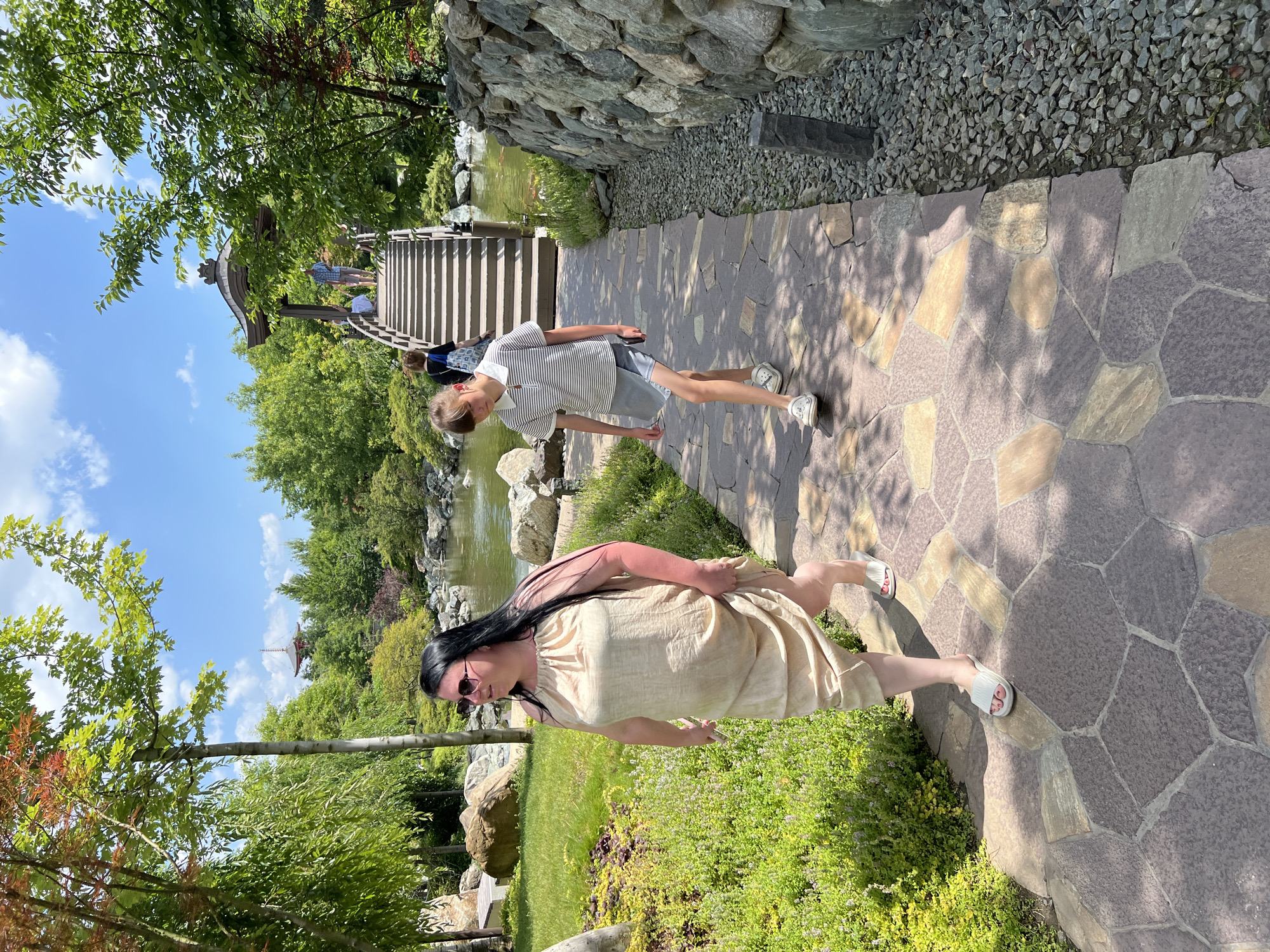 In this outdoor, full-shot image, two people are walking along a stone pathway in a park-like setting, possibly a botanical garden or an oriental garden. The pathway is made of large, irregular-shaped stones in shades of gray, beige, and purple, with patches of sunlight and shadow dappling the ground. To the left of the pathway, a body of water with lily pads and surrounding rocks is visible, leading to a wooden bridge with a distinctive pagoda-style roof. A set of wooden stairs ascends from the water's edge towards the bridge. Lush green trees and foliage line the water and the pathway, creating a vibrant natural backdrop. The sky is bright blue with scattered white clouds, suggesting a sunny day.
In the foreground, a woman with long dark hair and sunglasses is walking away from the camera, wearing a light-colored, sleeveless dress and white slide sandals. Her back is to the camera, and she appears to be looking down at the pathway. Further along the pathway and slightly ahead of her, a young person wearing a striped t-shirt and denim shorts is walking towards the camera, also in athletic shoes. This person is captured in motion, with one leg extended. The overall mood of the image is relaxed and leisurely, suggesting a pleasant day spent exploring an outdoor recreational area. The setting cues, such as the pagoda-style bridge and the carefully arranged stones and water, point towards an East Asian garden influence. The location is noted as Krasnodar, Russia. There is no visible text in the image.