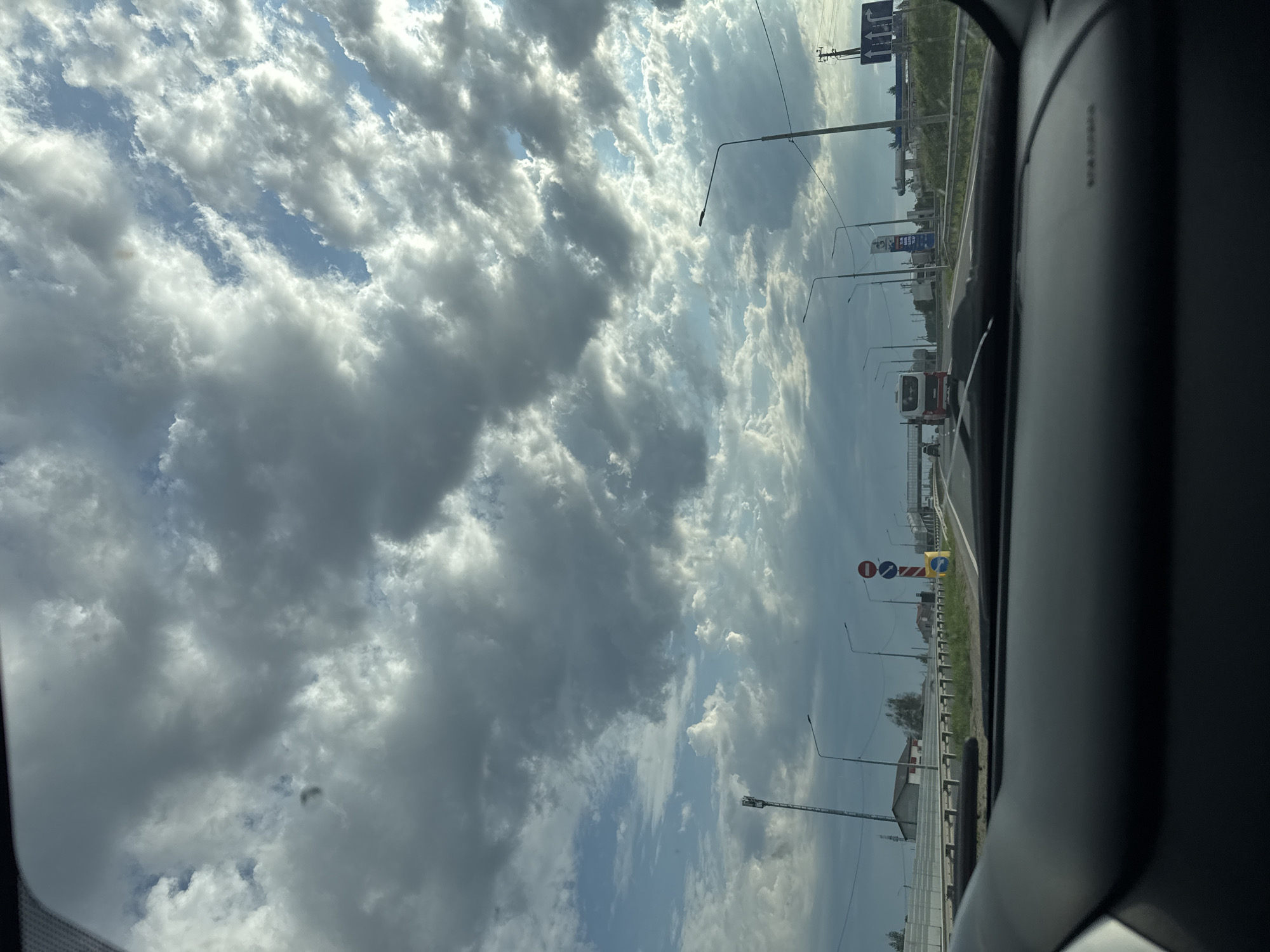 The image, taken from inside a vehicle, shows a view of the sky dominated by cumulus clouds. Through the vehicle's window, a roadside scene unfolds, featuring lampposts, road signs, and buildings. A red and white bus is visible in the distance. The location is identified as Усть-Сыны, Russia.