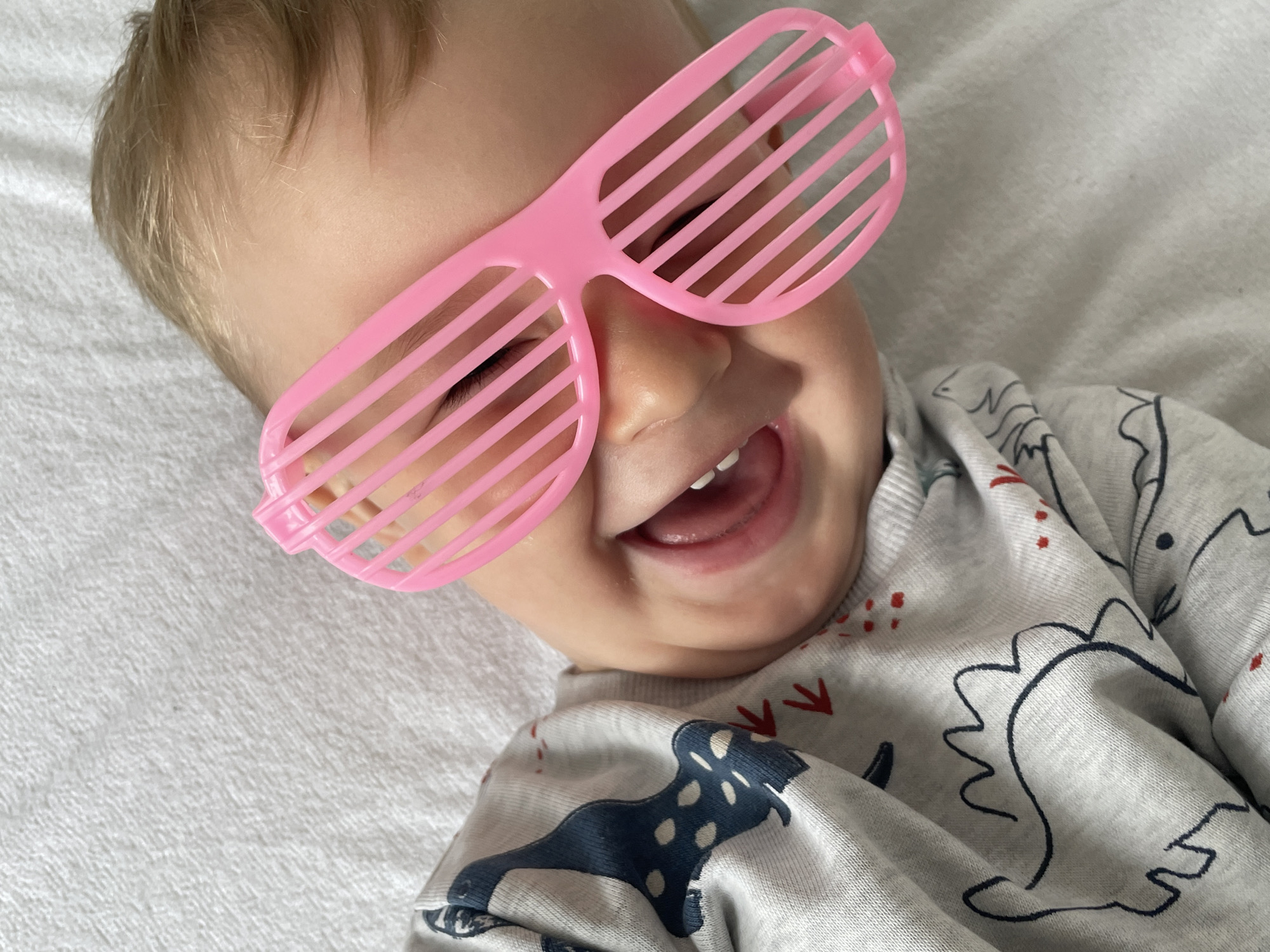 A baby is lying on a white textured surface, possibly a blanket or bed, with their head tilted. The baby is wearing a grey long-sleeved shirt with a dark blue dinosaur pattern and red accents. They are also wearing pink shutter-style glasses that partially cover their face. The baby's mouth is open, revealing their teeth and tongue, and their eyes are closed or obscured by the glasses. The lighting is soft, suggesting an indoor setting. There is no discernible location in the city of Nidda, Germany.